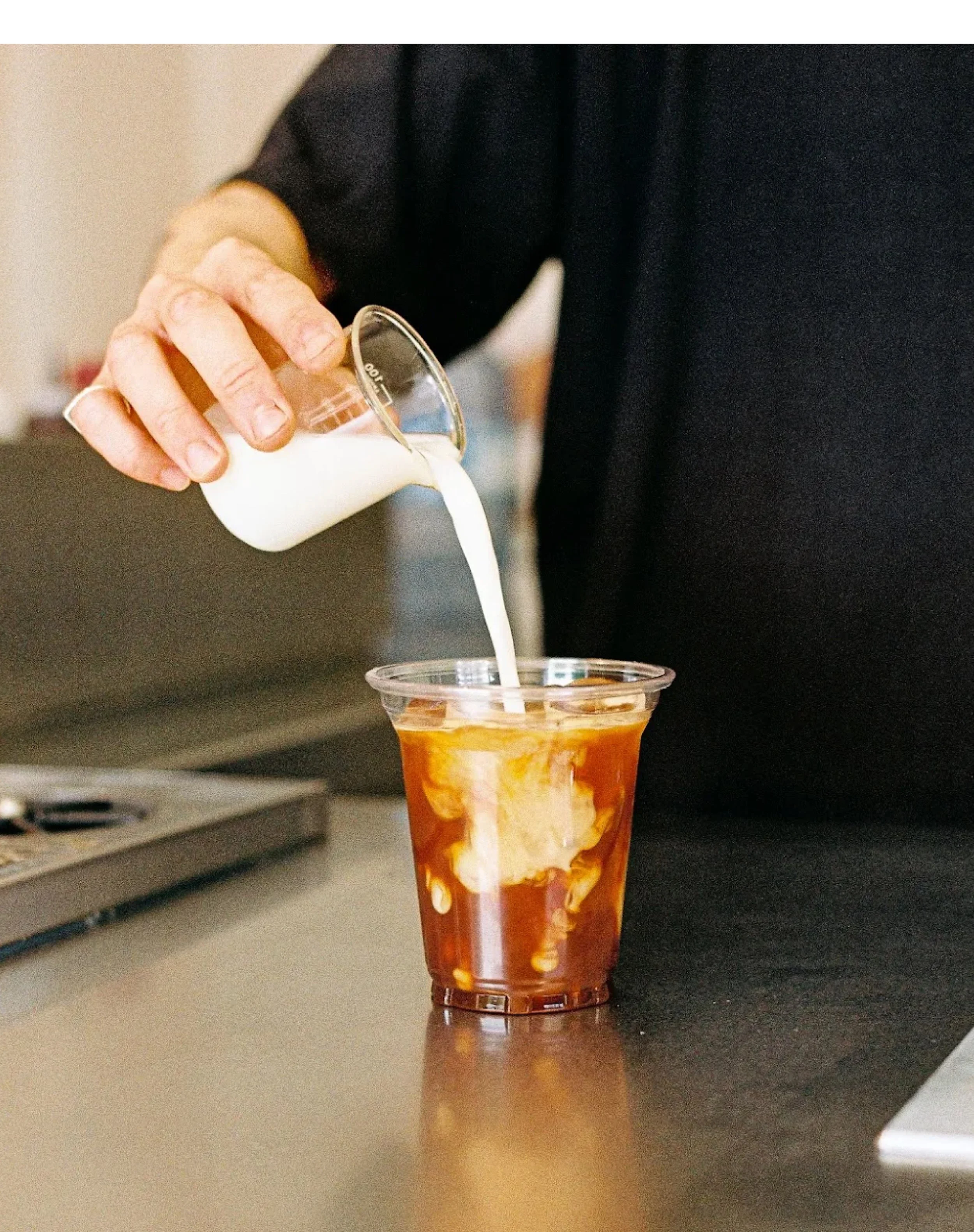 Person pouring milk into a caramel-colored beverage with swirls in a clear plastic cup, on a kitchen counter.