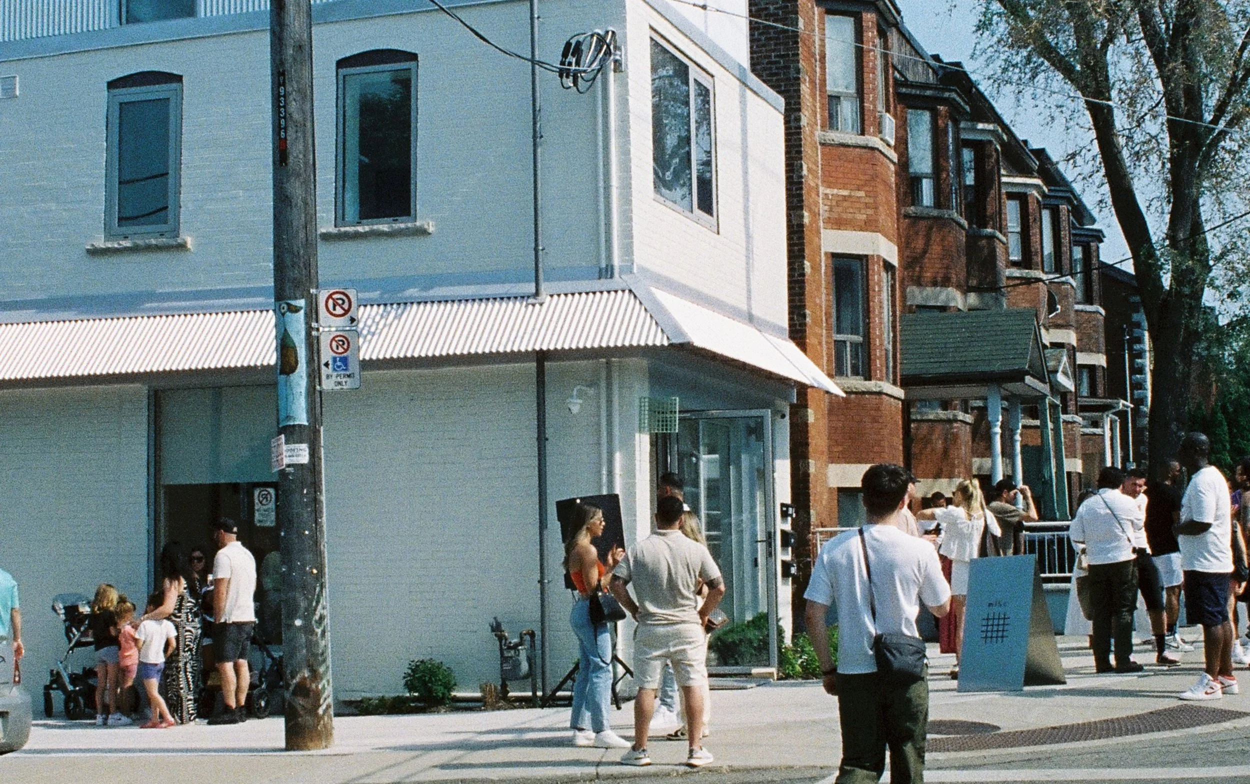 Crowd of people walking along a city sidewalk in front of buildings with storefronts and large windows, under a clear sky.