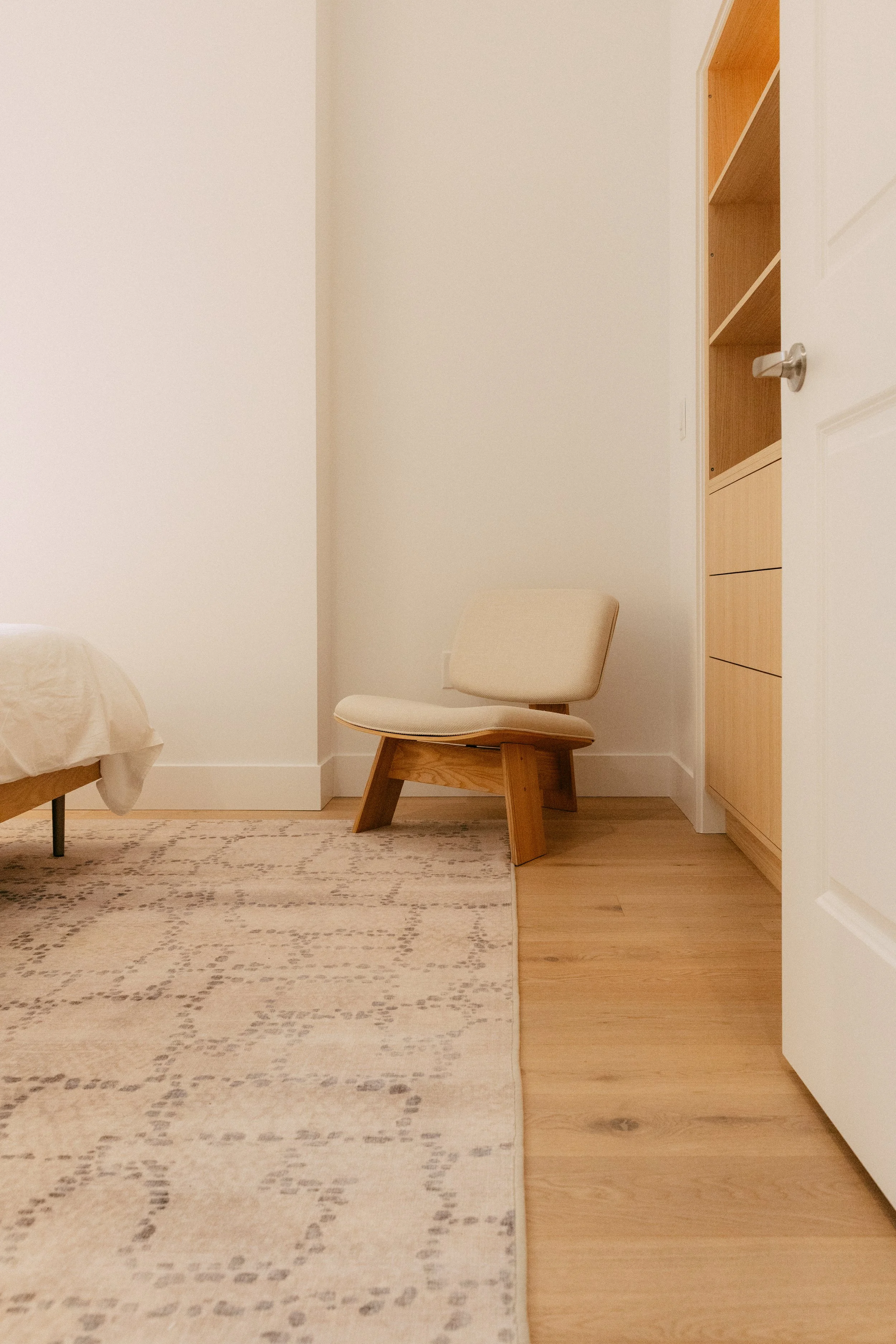 A corner of a room with a beige chair, a stool with wooden legs, and a built-in wooden shelf with three open compartments.