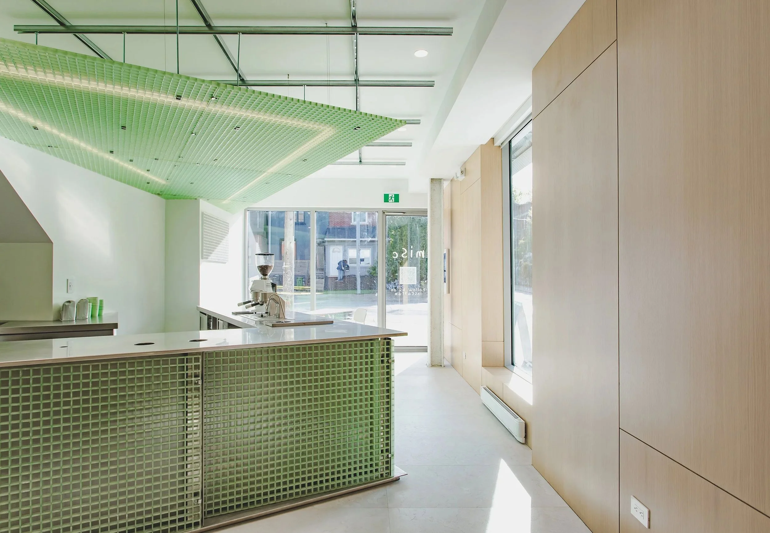Modern interior space with string green ceiling installation, light wood wall panels, large glass windows, and a countertop with coffee equipment.