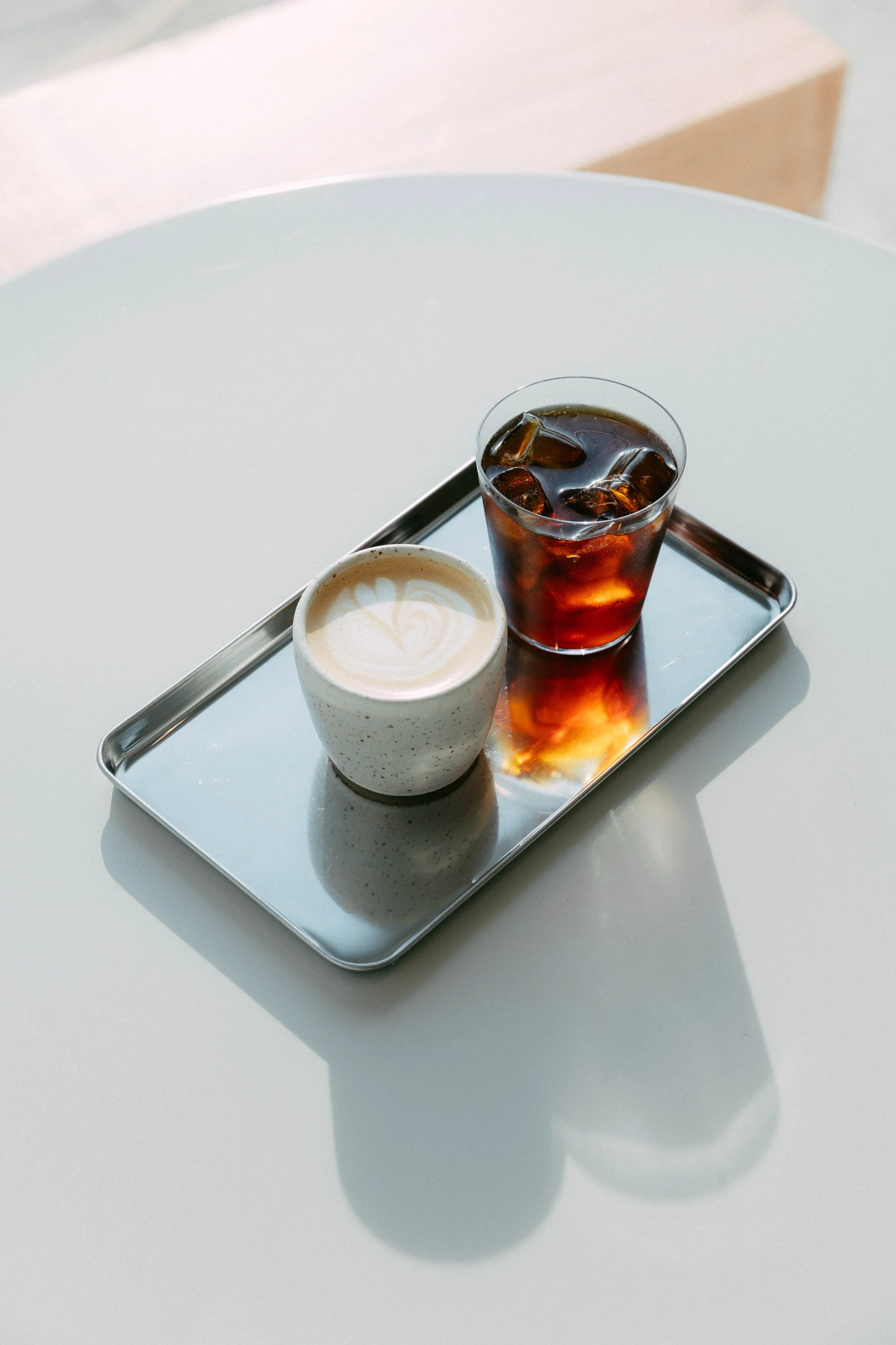A tray with a cup of latte art coffee and a glass of iced cola on a white surface.