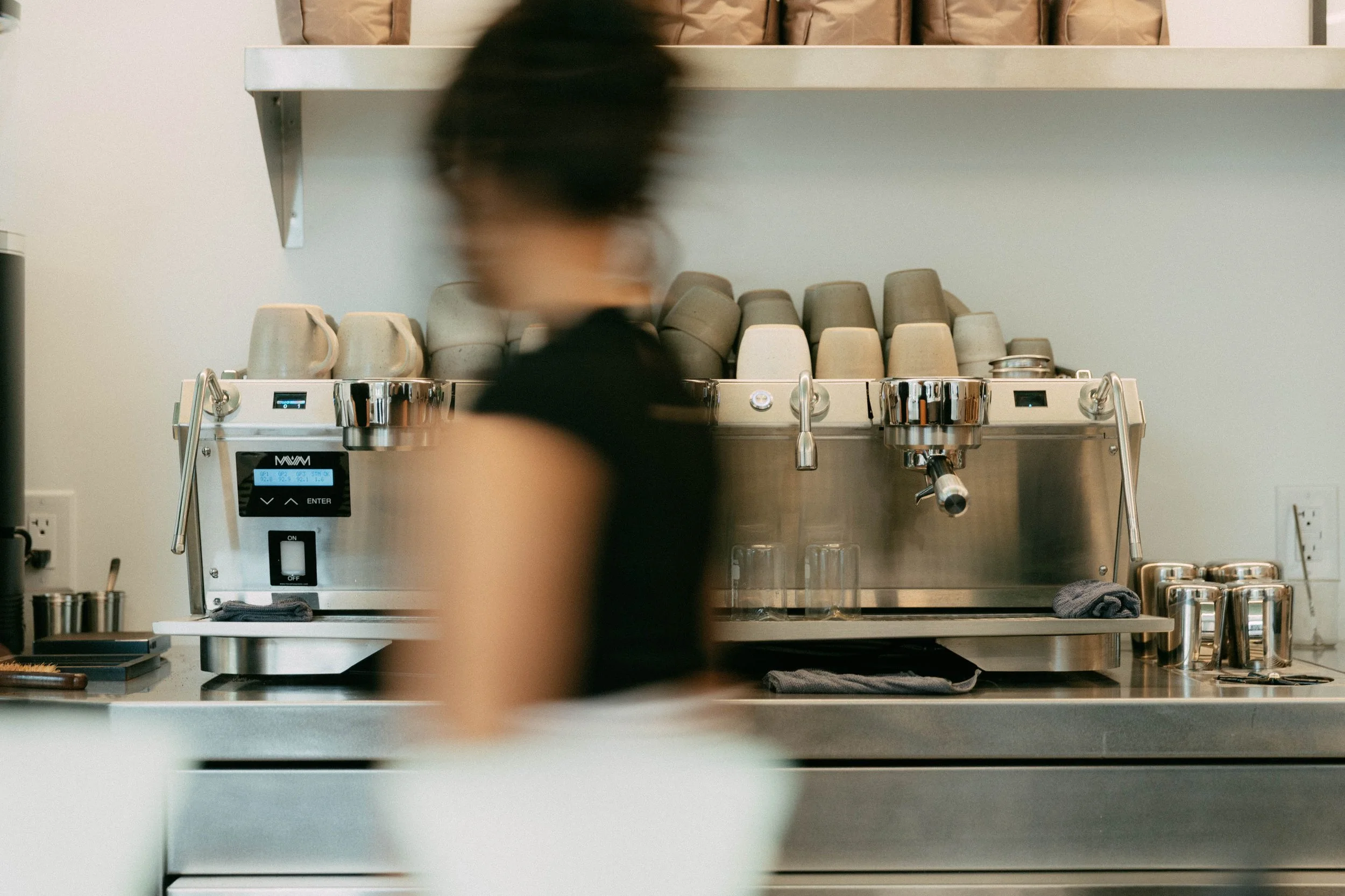 A blurred person working behind a professional espresso machine with cups and steam wands, in a modern café or coffee shop.