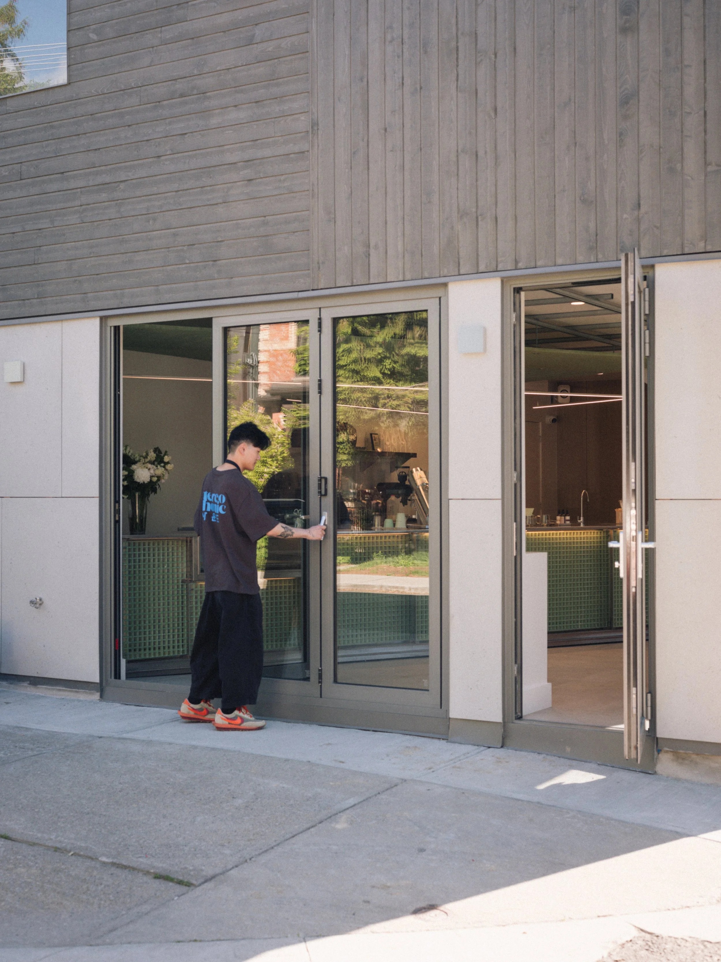 A person with black hair, wearing a black shirt and orange sneakers, standing outside a modern building with large glass doors and wooden panel siding, holding the door open.