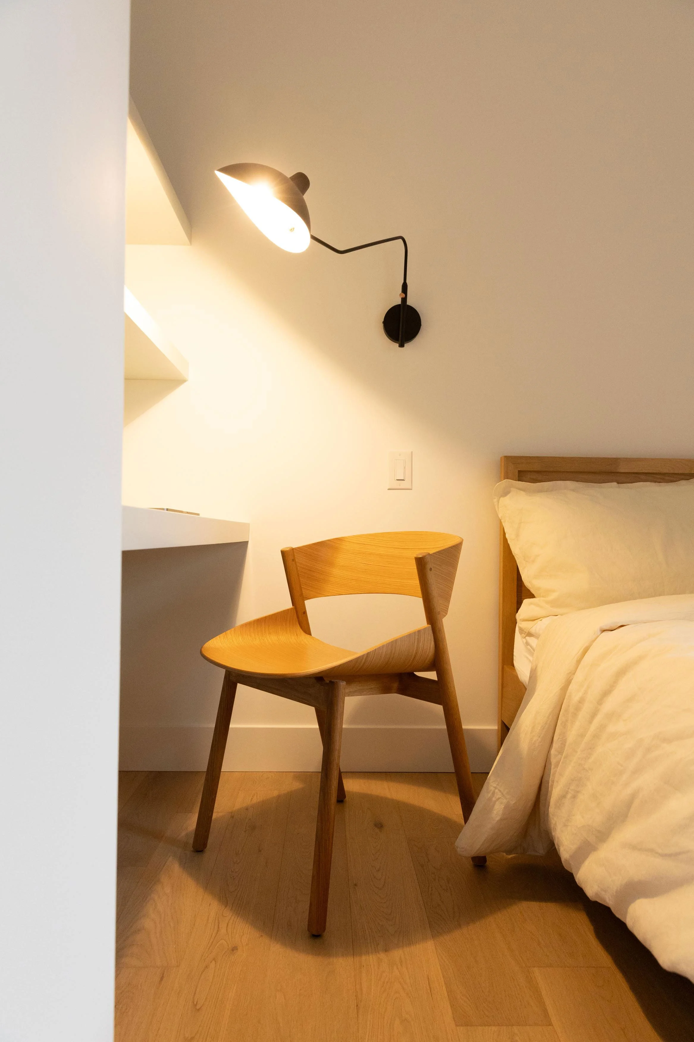 A bedroom scene with a bed, wooden chair, wall lamp, and a side table, with warm lighting and minimal decor.