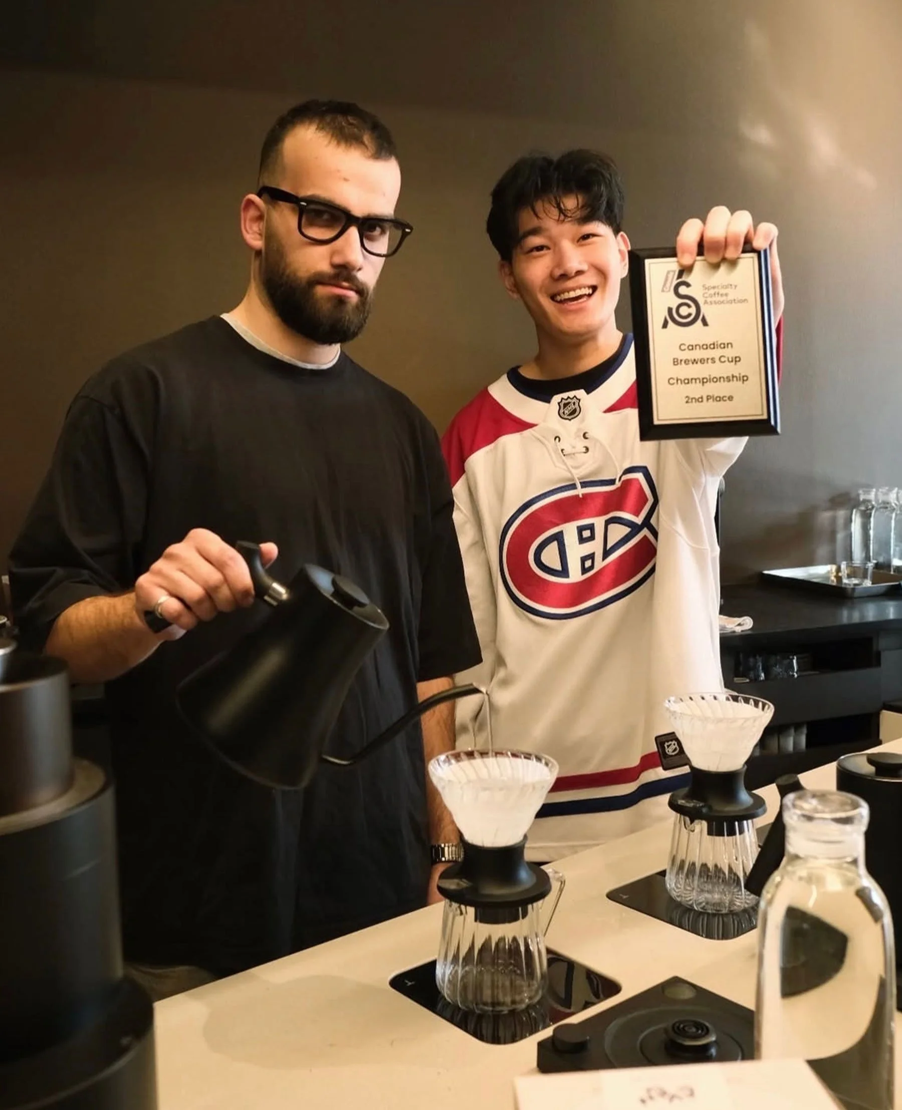 Two men standing behind a kitchen counter, one holding a plaque that reads 'Canadian Brewers Cup Championship 2nd Place.' The man on the left has a beard and glasses, and is wearing a black shirt, while the man on the right is smiling and wearing a hockey jersey. There are coffee-making equipment and glasses on the counter.