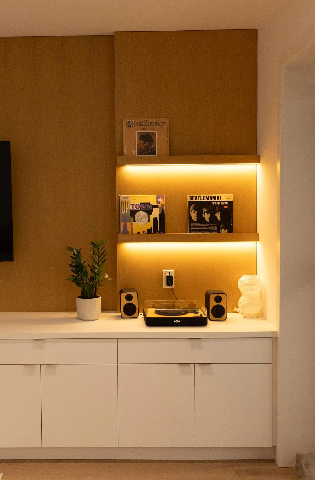 A modern white cabinet with multiple drawers, a potted plant, a turntable, and two speakers on a white surface, with a wallpapered wall behind featuring framed John Lennon artwork, vinyl records, and a lamp with a wavy design.