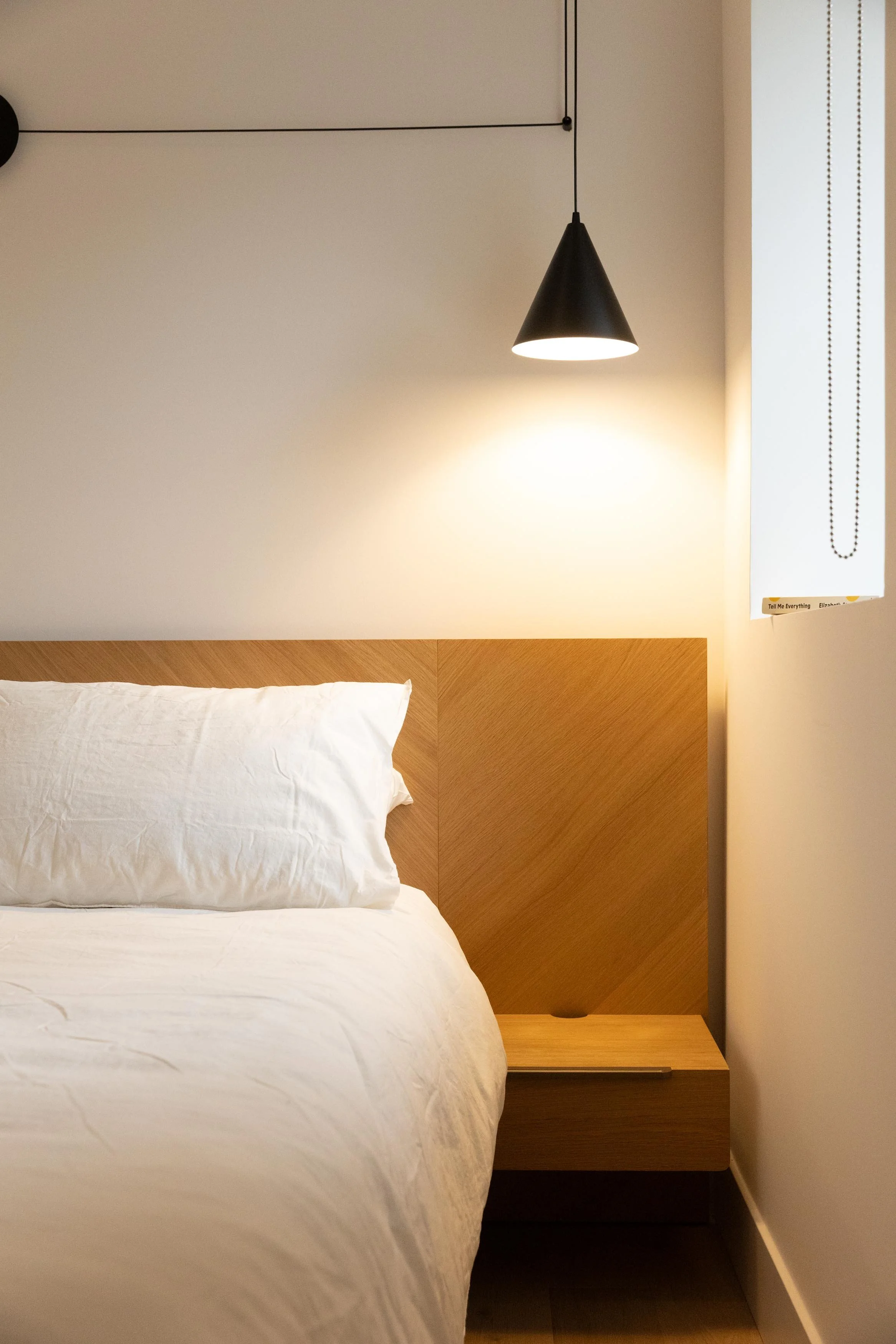 A neatly made white bed with a white pillow against a light-colored wall. Above the bed, a black cone-shaped pendant light hangs from a black cord. To the right, there is a window with a white roller shade and a small piece of yellow paper or card visible in the bottom right corner of the shade.