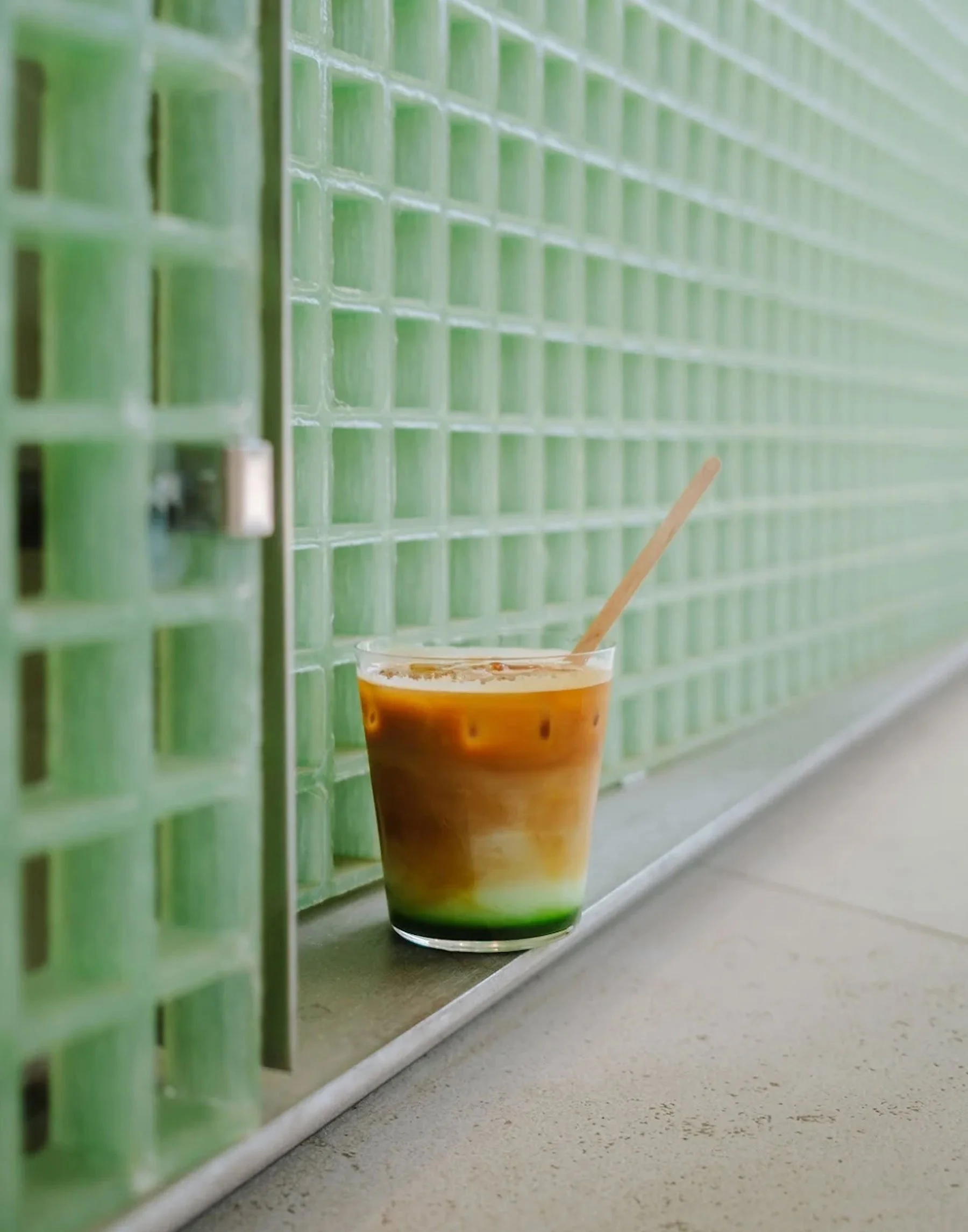 Close-up of a layered drink in a clear glass placed on a concrete ledge, with a green tiled wall in the background.