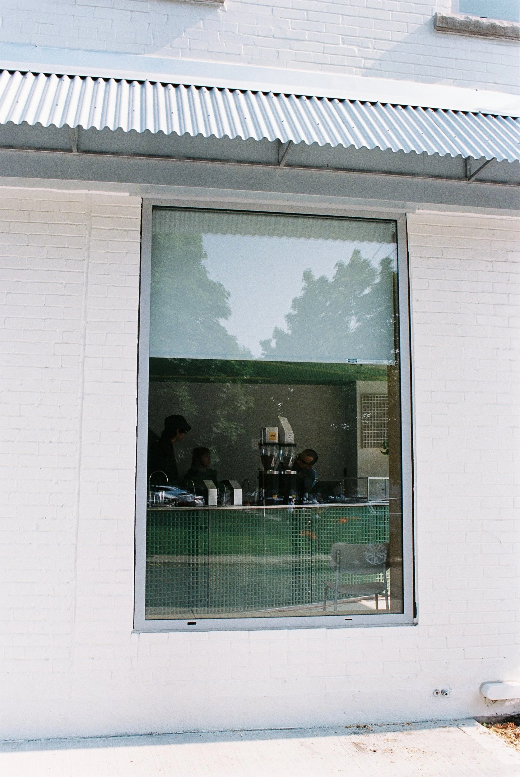 The image shows the exterior of a building with a large glass window. Inside, there are people sitting at a table, with some items like a coffee machine visible on the counter. The building has a white brick wall, a metal awning, and a reflection of trees in the window.