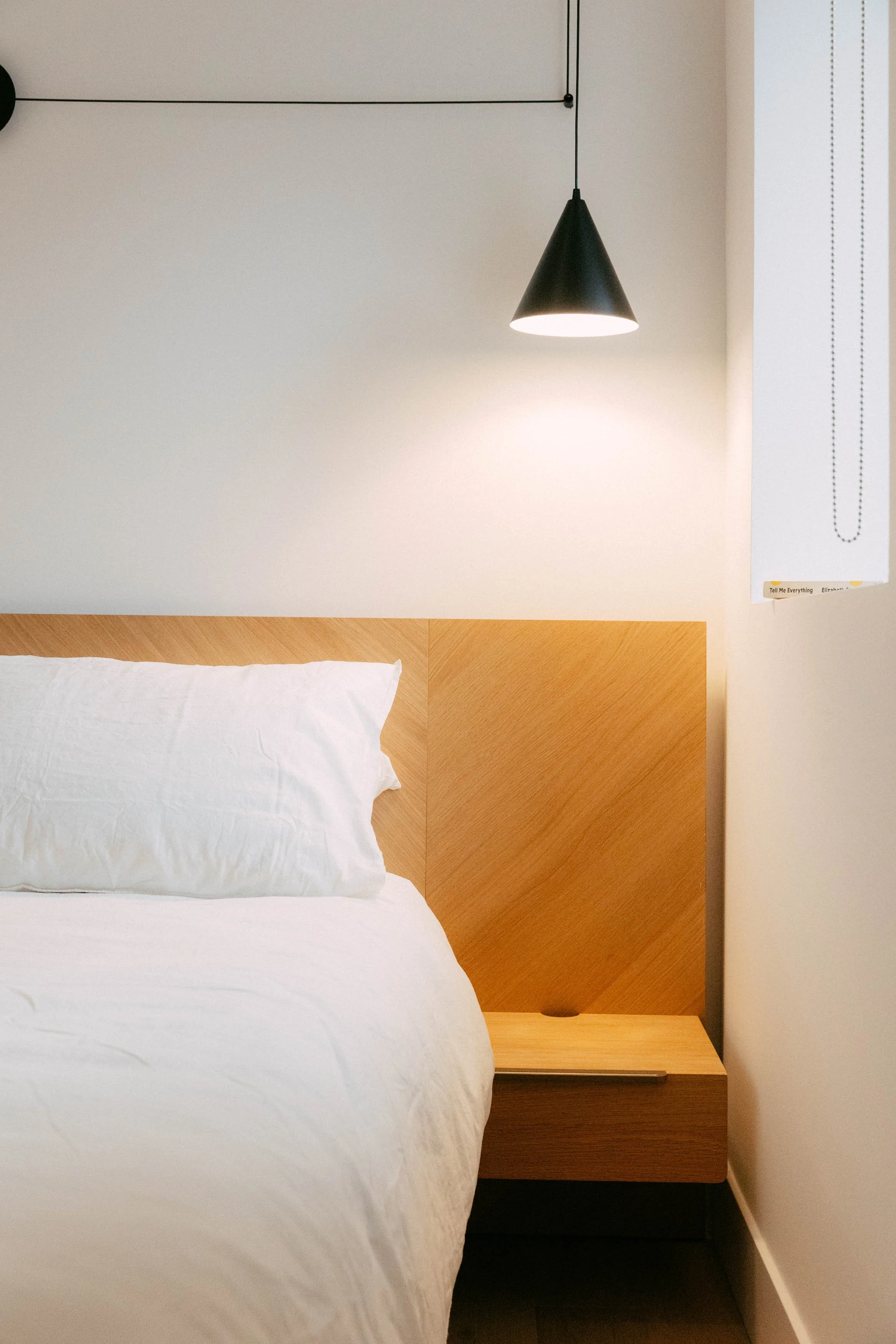A neatly made bed with white sheets and pillow in a modern minimalistic bedroom. A wooden headboard and nightstand are visible, with a black hanging cone-shaped pendant light above.