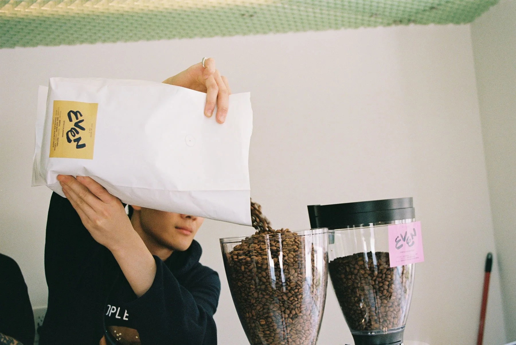Person pouring coffee beans from a white bag into a glass container at a coffee shop.