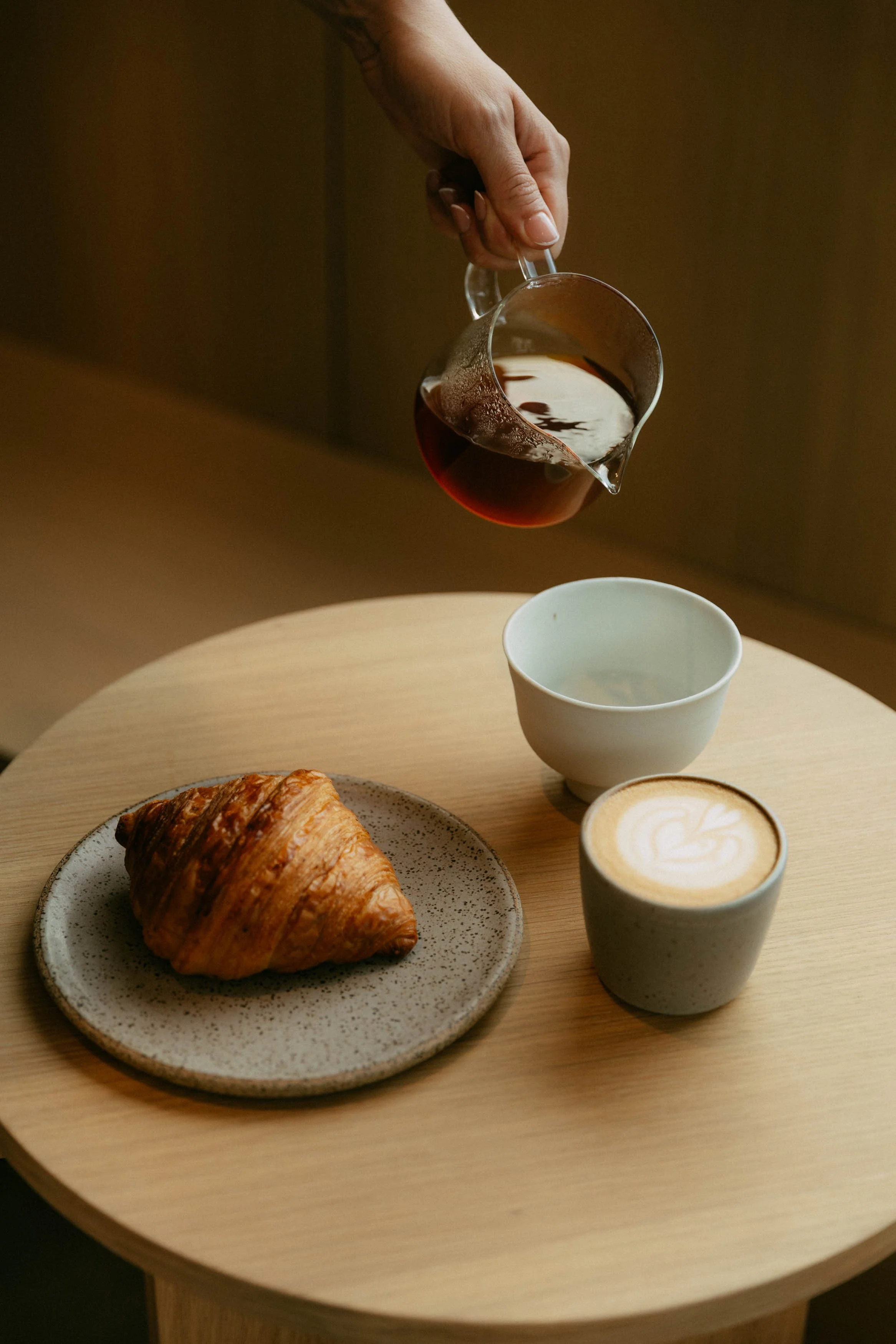 A table with a croissant on a speckled plate, a cup of coffee with latte art, two cups of hot beverages, a glass of red wine, and a white cup on a wooden table.