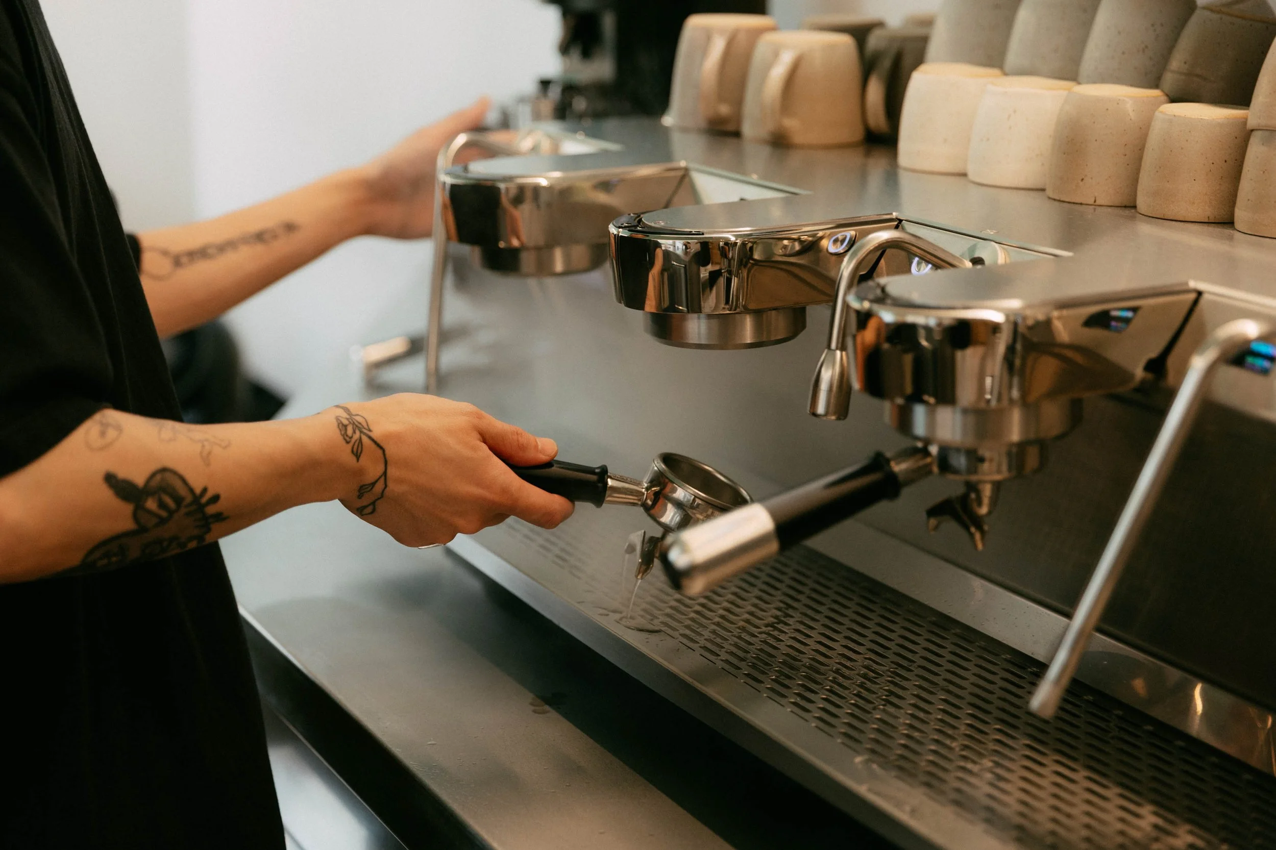 A barista with tattoos on their arms preparing coffee at an espresso machine in a coffee shop.