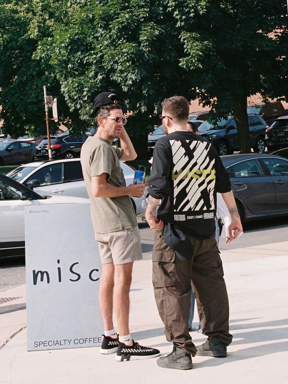 Two men standing and talking on a sidewalk with parked cars and green trees in the background. One man is wearing a beige shirt, shorts, a black baseball cap, and checkered sneakers. The other man is wearing a black and white graphic sweatshirt and baggy pants.