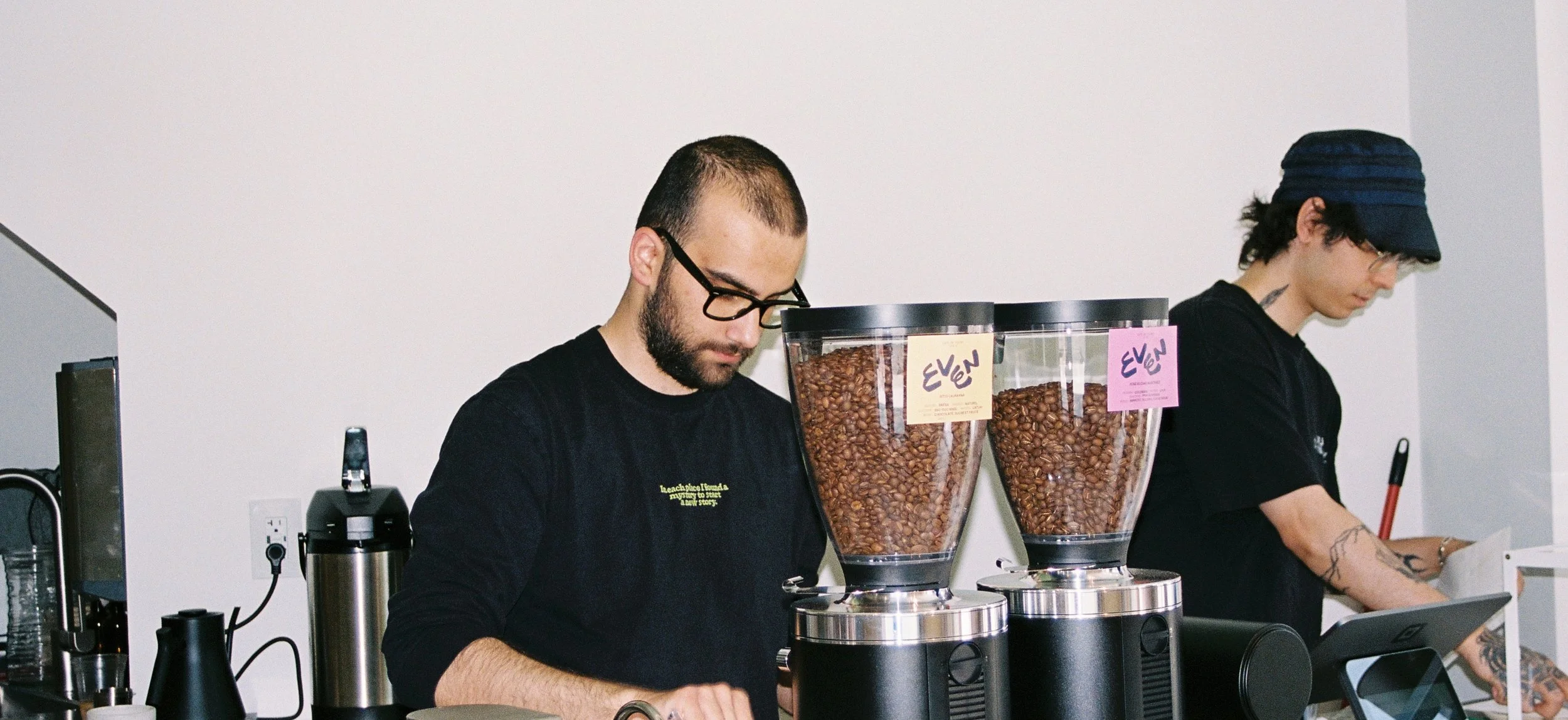 Two baristas working behind a coffee counter. The barista in the foreground has a beard and glasses and is preparing coffee. The barista in the background has tattoos and is wearing a dark hat. There are two large containers of coffee beans with labe