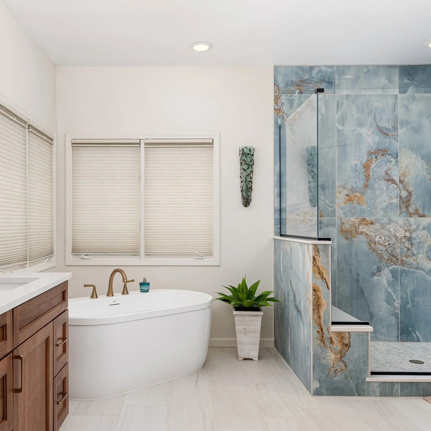 Modern bathroom with white walls, large windows with blinds, a freestanding bathtub with brass fixtures, a small plant in a white pot, and a walk-in shower with blue and brown marble tiles.
