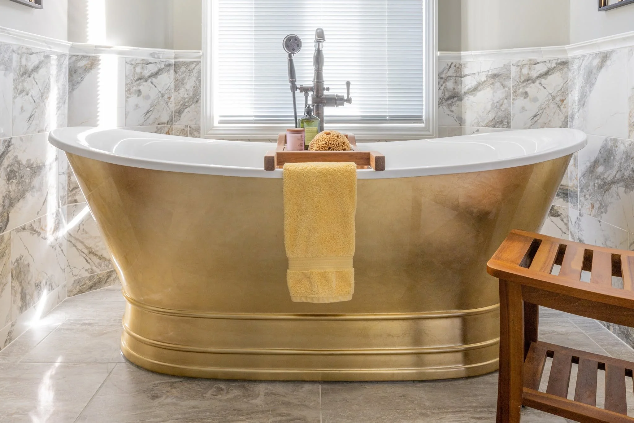 A clawfoot bathtub with a yellow towel hanging over its edge, with bottles and a sponge on a tray, situated in a bright bathroom with marble tiled walls and a window with blinds.