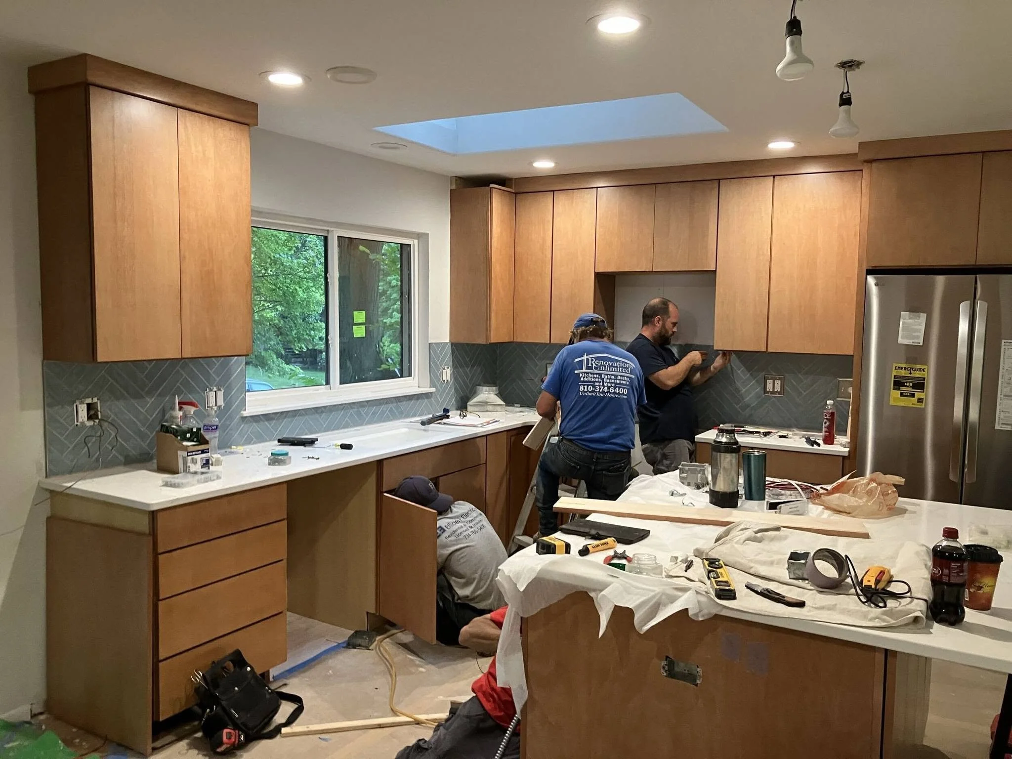 Two workers are installing kitchen cabinets. One is wearing a blue shirt with text on the back, and the other is in a dark shirt. The kitchen has wooden cabinets, a window showing green trees outside, and various tools and materials on the counter and table.