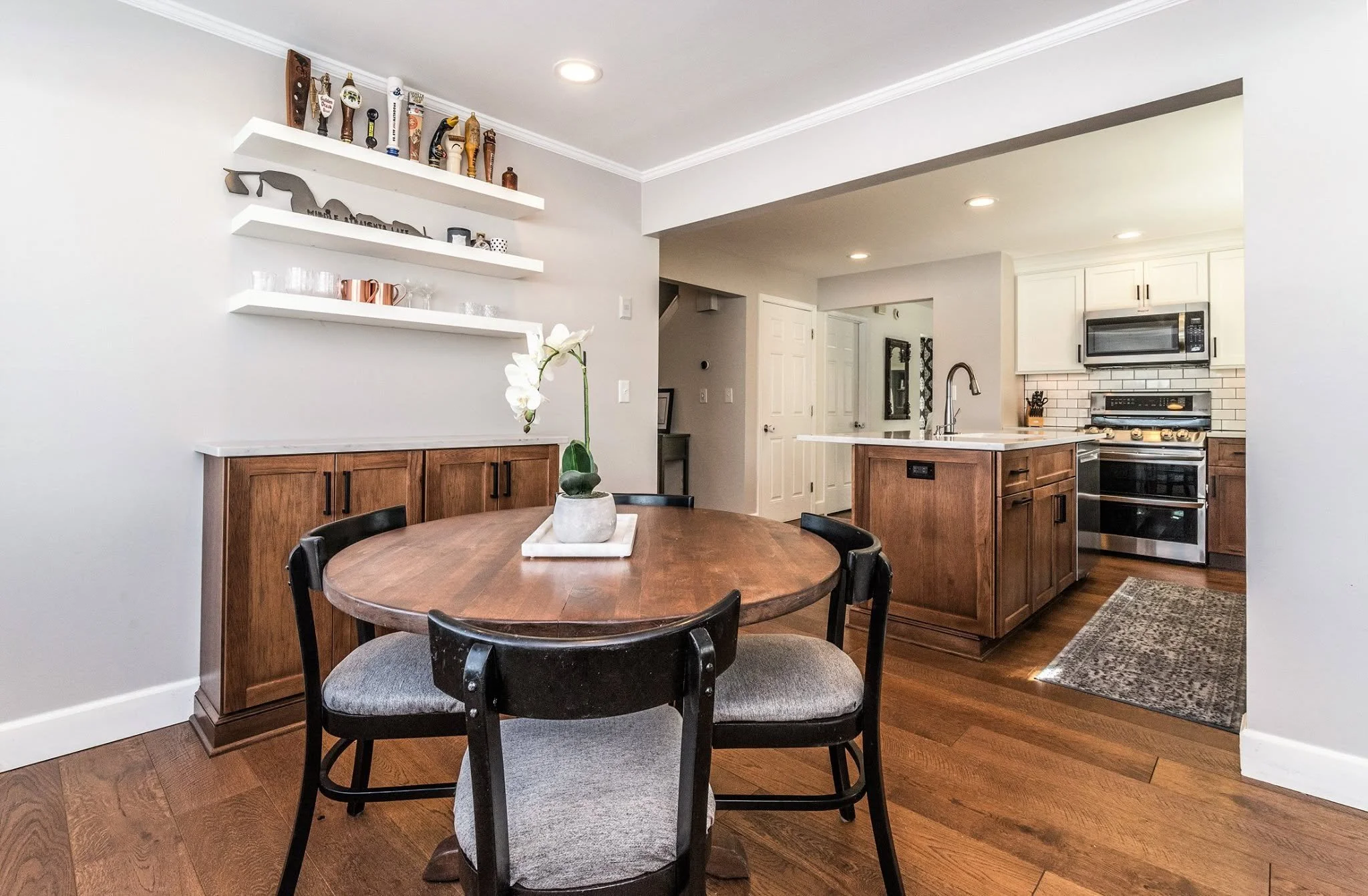 A cozy kitchen with a round wooden dining table, black chairs, a vase with white orchids, white floating shelves with decorations, and stainless steel appliances.