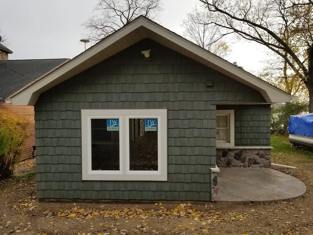 Small house with green shingle siding, white-framed window, and stone foundation, with a small concrete patio and leaf-covered ground in the yard.