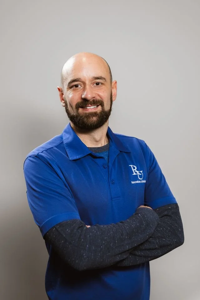 A man with a beard, smiling, wearing a blue polo shirt with a logo, standing with arms crossed against a plain background.