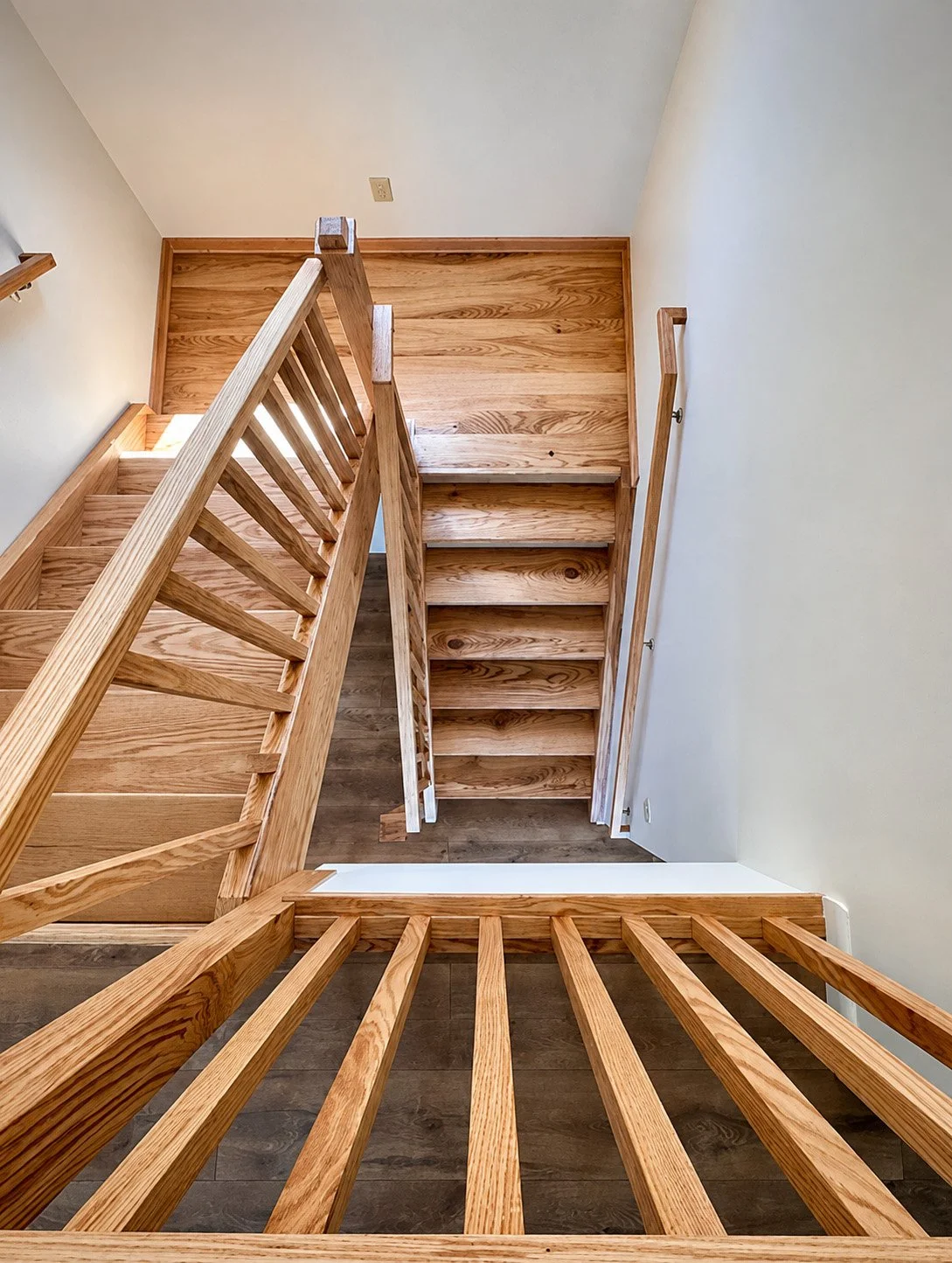 A wooden staircase viewed from above, showing multiple steps and a railing on the left side, leading down to a lower floor with brown wooden flooring and white walls.