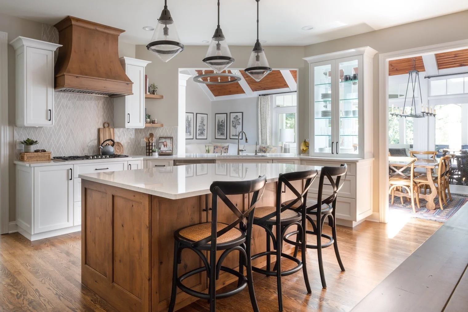 Modern kitchen with white cabinets, wooden island, hanging pendant lights, and adjacent dining area with wooden table and chairs, bright and airy with large windows and wooden ceiling accents.