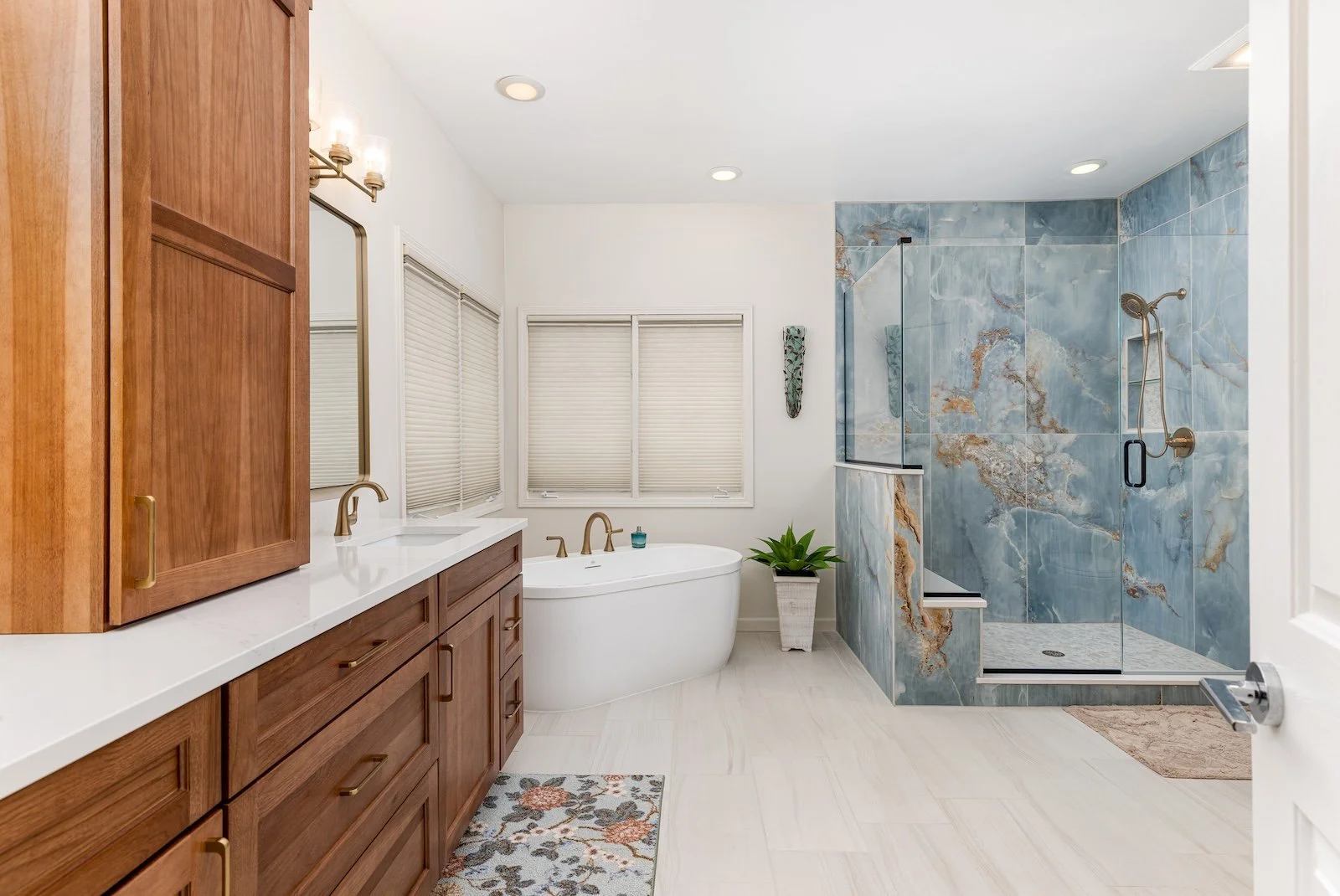 Bathroom with wooden vanity, white countertops, double sinks, and a large mirror. Includes a freestanding bathtub with a plant nearby, and a glass-enclosed shower with blue and brown marble tiles.