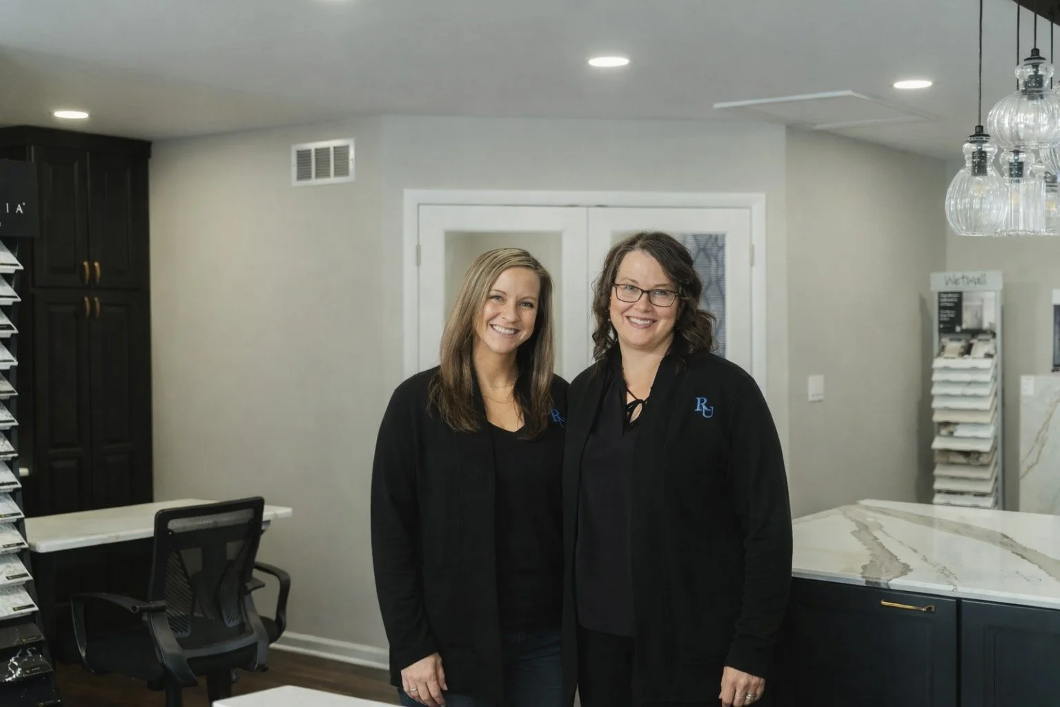 Two women smiling inside a modern kitchen, standing in front of a white door, with shelves displaying material samples in the background.