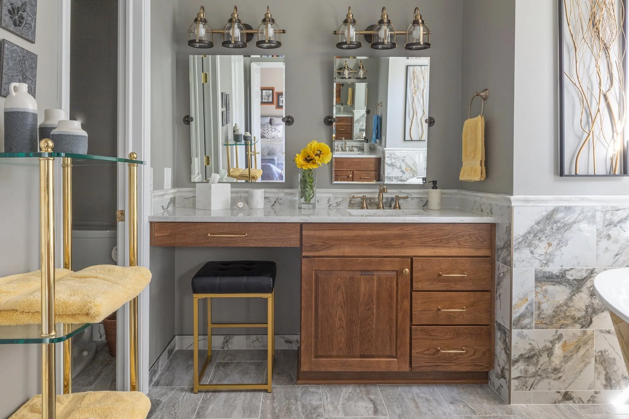 A bathroom vanity with a marble countertop, wooden cabinet drawers, a small black stool, a vase with yellow flowers, and a mirror. To the left, a glass shelf with decorative vases, and to the right, a yellow towel on a hook and a decorative wall art piece. The bathroom has gray walls and marble flooring.