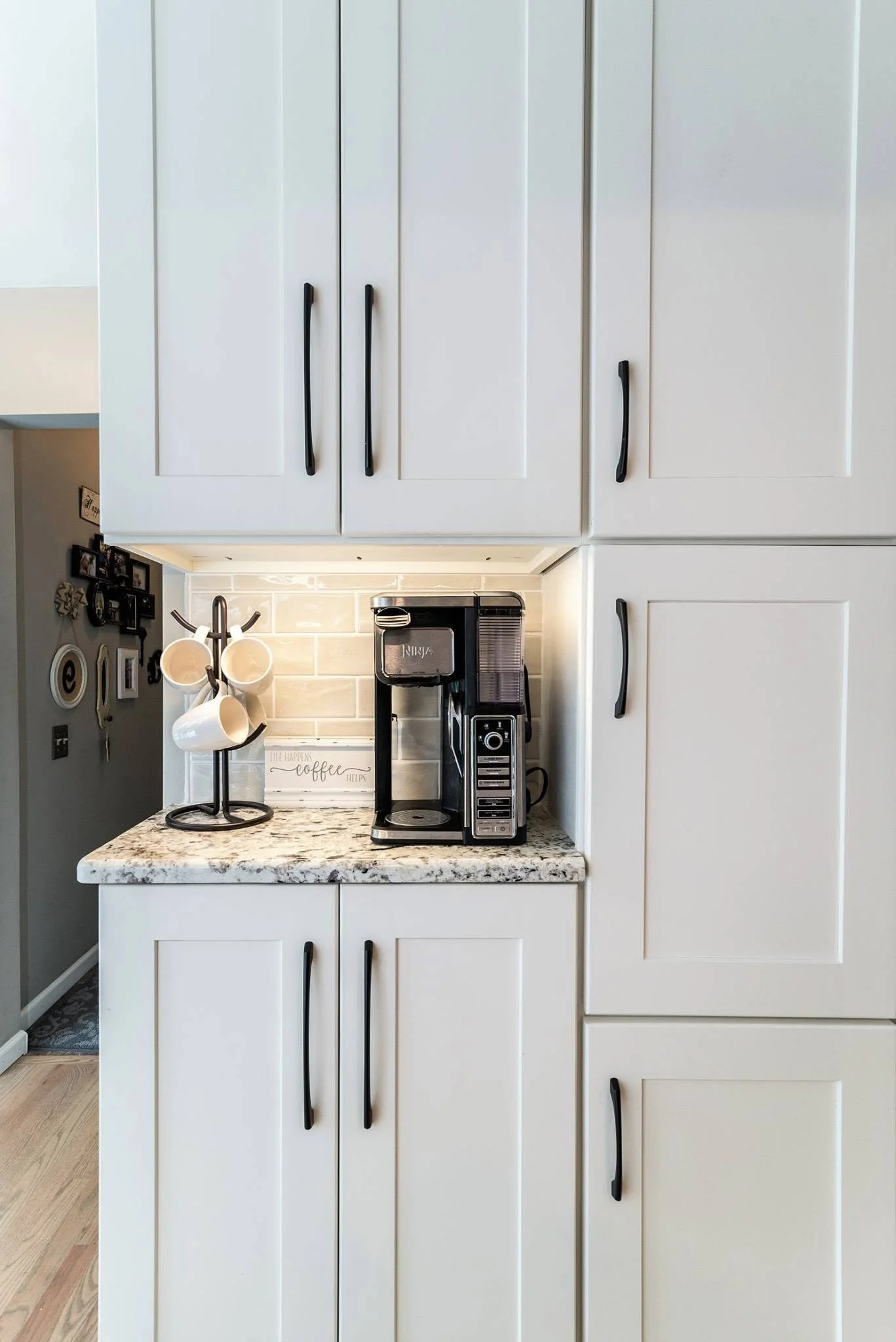 White kitchen cabinets with black handles, a coffee maker on granite countertop, hanging mugs, and a small framed quote, with a partial view of a wall with photographs and decorations.