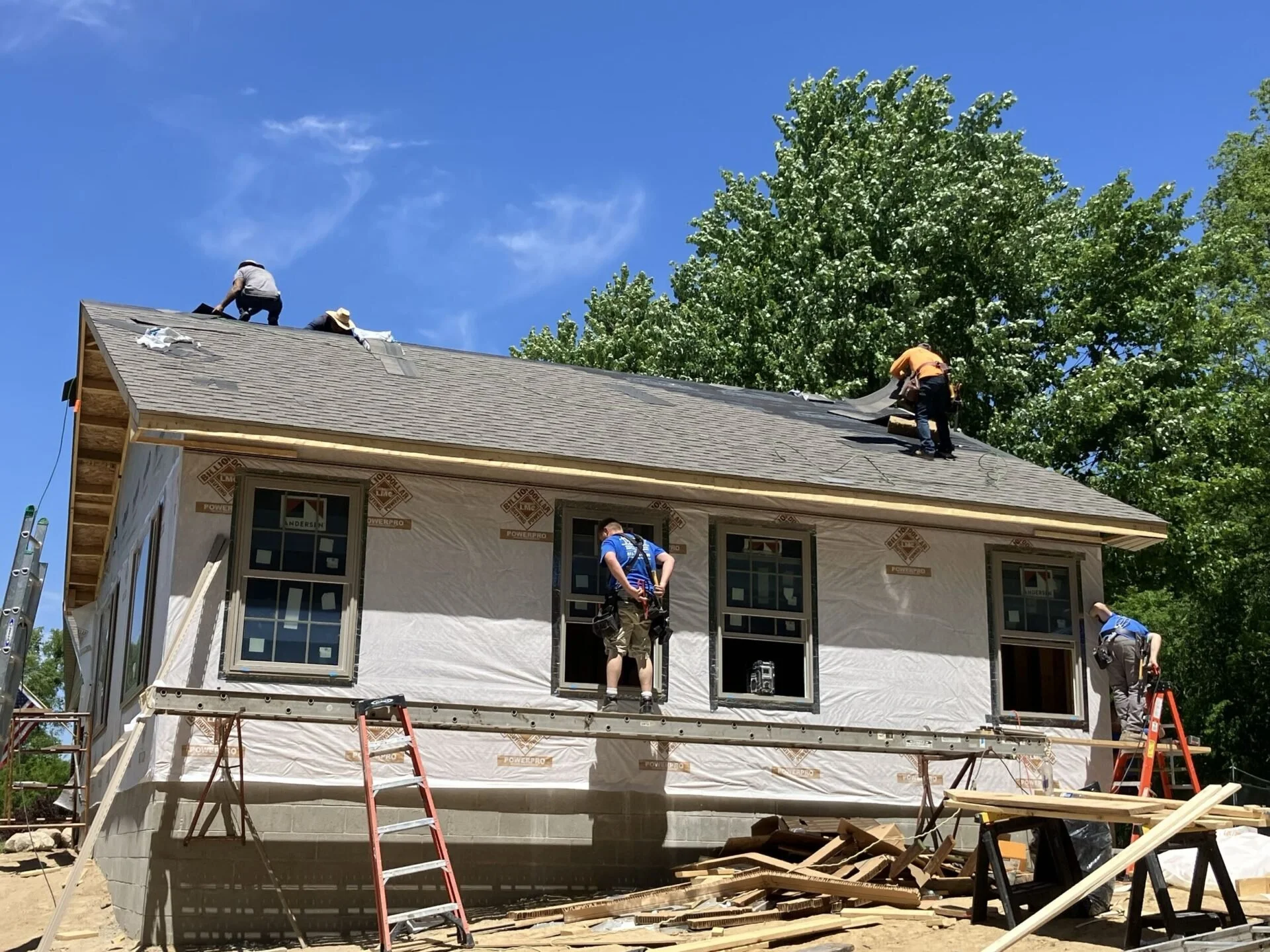 Construction workers installing roofing on a house, with a tree and blue sky in the background.
