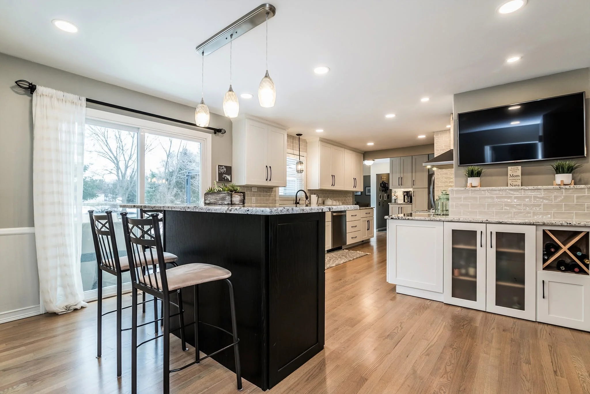 Modern open-concept kitchen and dining area with white cabinets, granite countertops, a tiled backsplash, large window with curtains, and a black kitchen island with bar stools. Recessed ceiling lights and a wall-mounted TV are also visible.