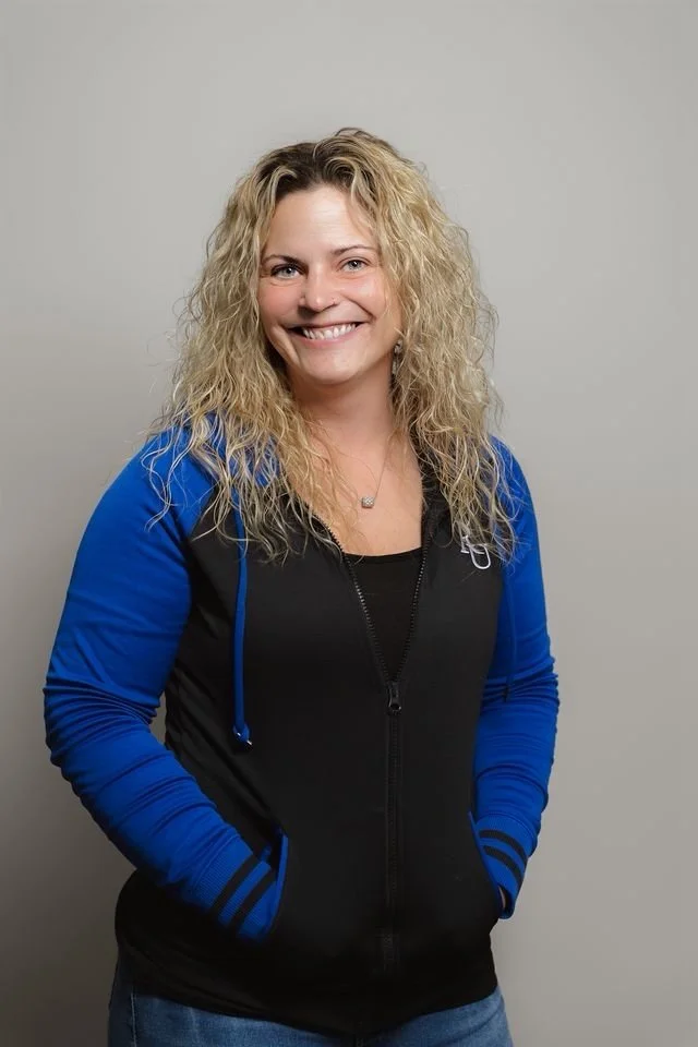 A smiling woman with curly blonde hair wearing a black and blue jacket and blue jeans standing against a plain light gray background.