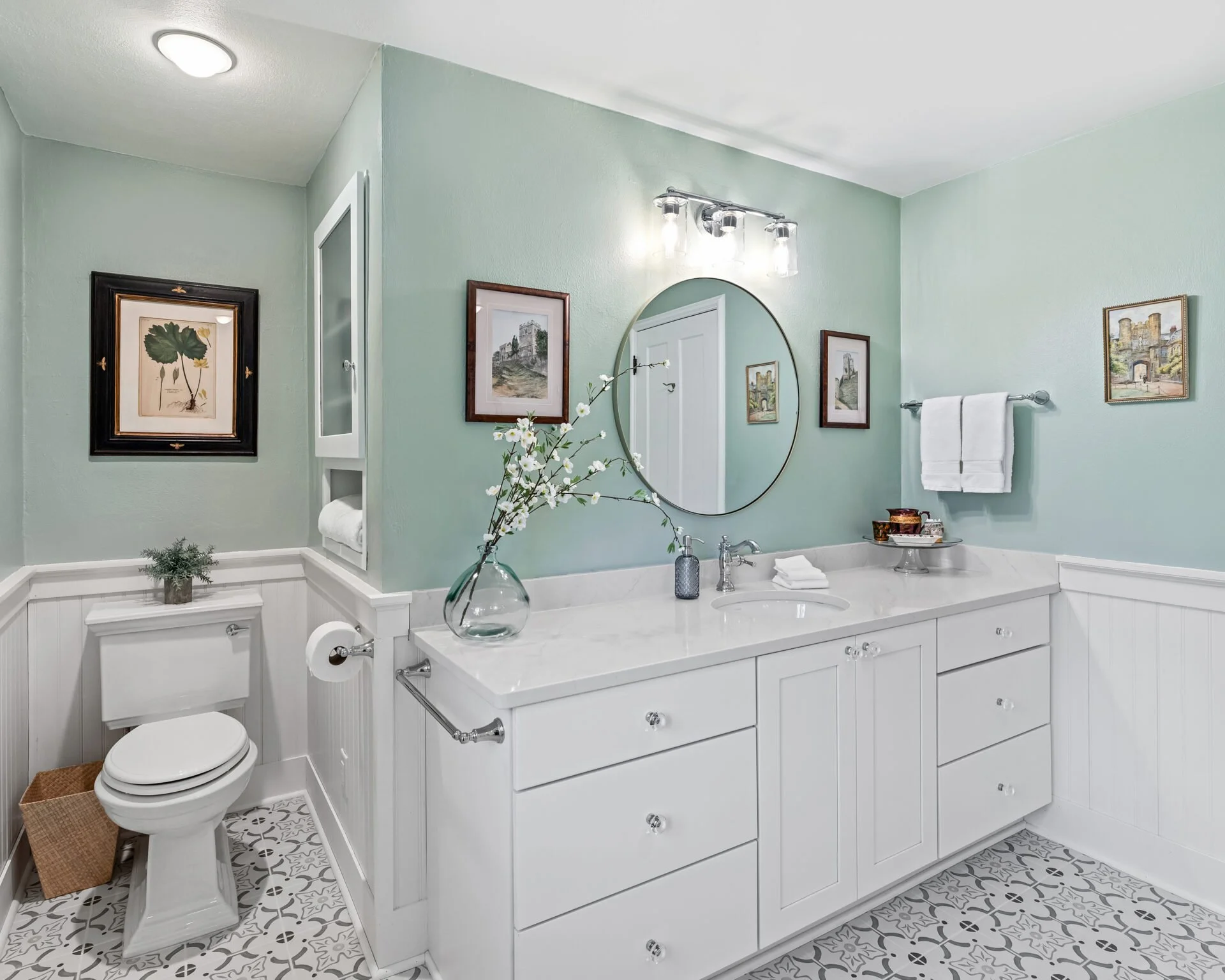 Modern bathroom with light green walls, white cabinetry, marble countertop, round mirror, decorative framed artwork, white towels on a chrome towel bar, vase with white flowers, patterned floor tiles, and ceiling light fixture.