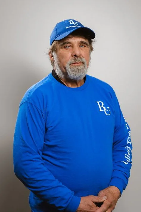 An older man with gray hair and beard wearing a blue baseball cap and matching blue sweatshirt with white emblem, standing against a plain background.