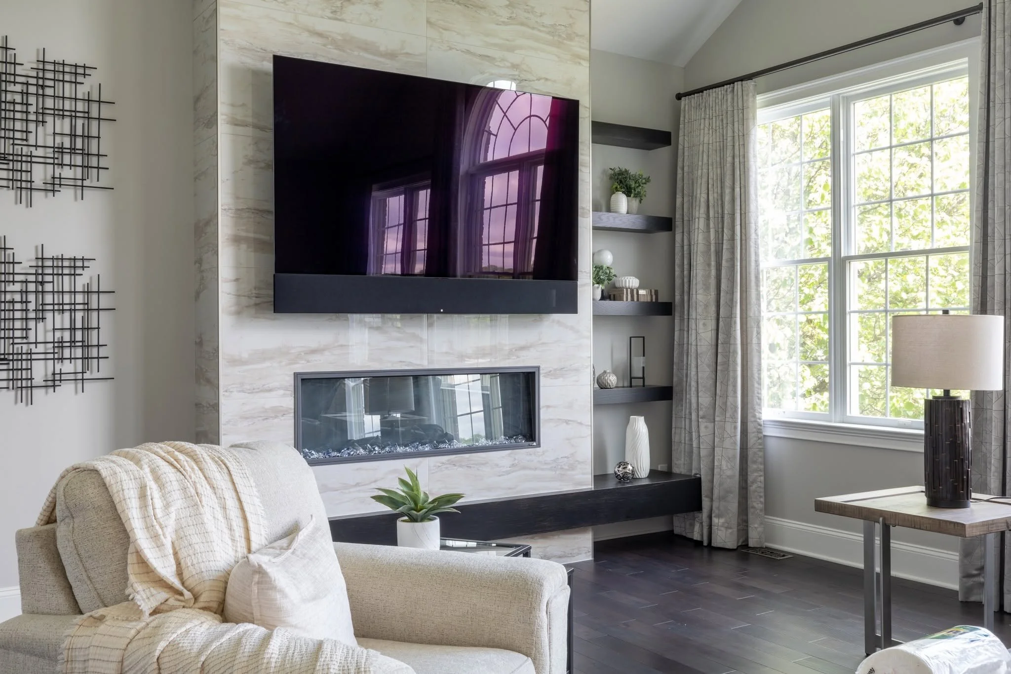Living room with beige sofa, cream throw blanket, black metal wall art, marble fireplace, wall-mounted TV reflecting window, corner shelves with decorative items, large window with patterned curtains, beige lampshade, wooden table, potted plant.