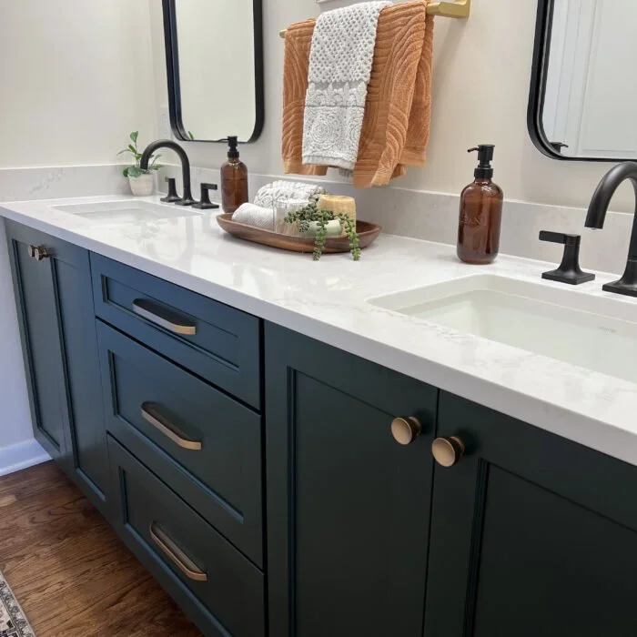 Bathroom vanity with a white countertop, dark blue cabinets with gold handles, a black sink faucet, and decor including a tray with towels, a potted plant, and two amber soap dispensers. Two mirrors are above the countertop.