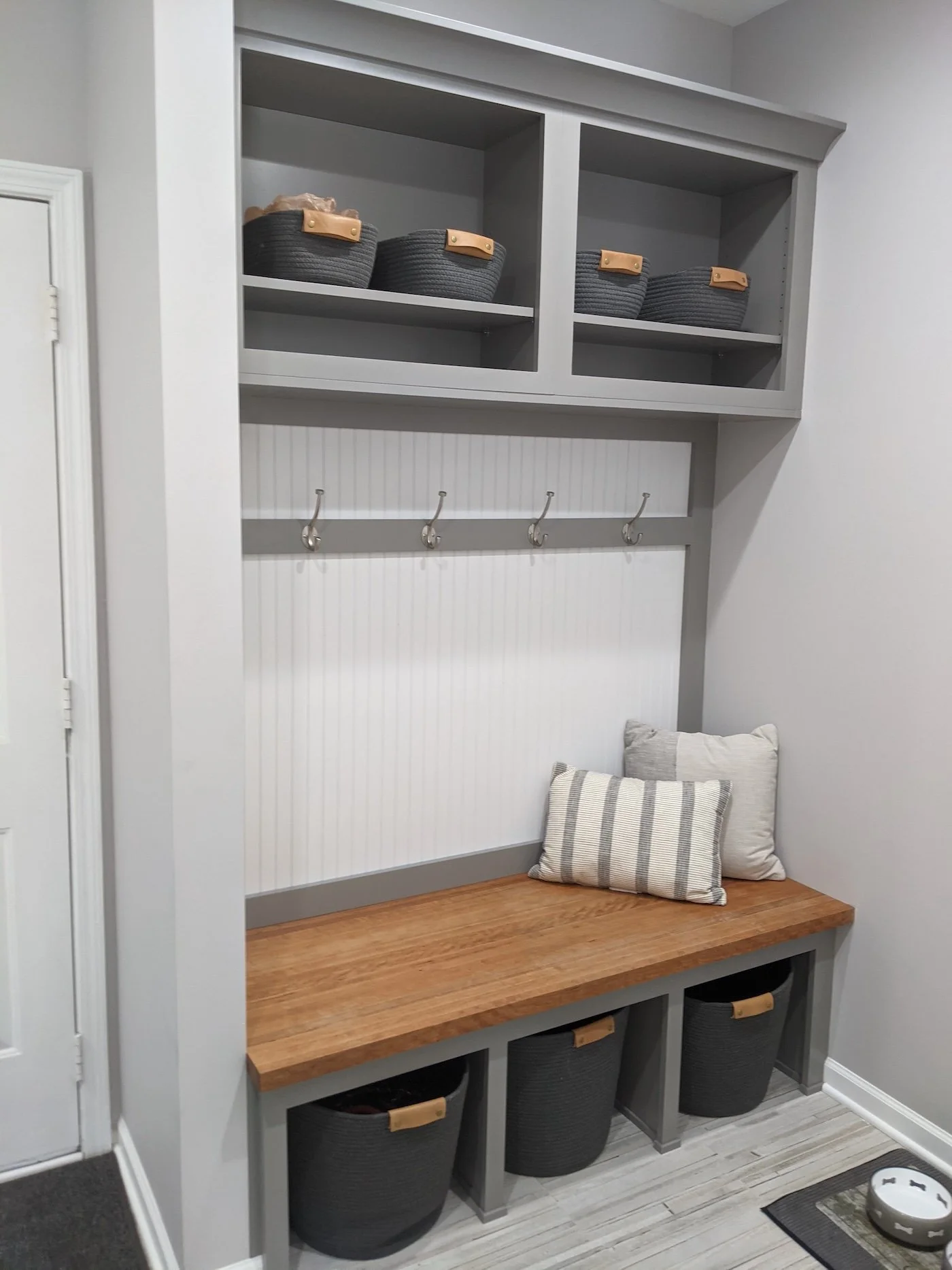 Gray and white mudroom or entryway organization bench with storage and baskets, pillows on the bench, and a rug on the floor.