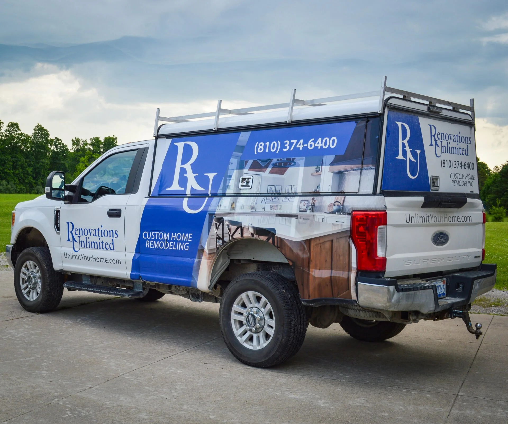 White truck with advertising wrap for Renovations Unlimited, showing contact phone number, website, and services like custom home remodeling, parked on a concrete surface with green trees and a cloudy sky in the background.
