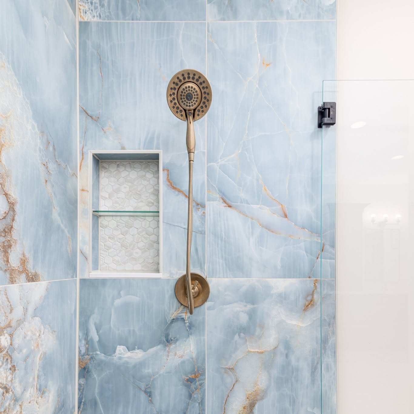 Close-up of a shower area with blue marble-patterned tiles, a round metallic showerhead, a built-in hexagonal tile shower niche, and a glass shower door with a black hinge.