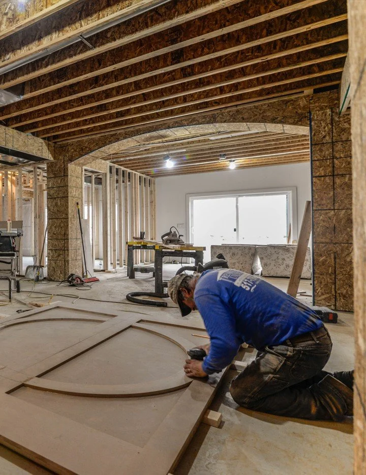 Construction worker kneeling on the floor, installing or working on a large wooden frame or decorative piece in a partially finished home interior with exposed ceiling beams and a large window in the background.