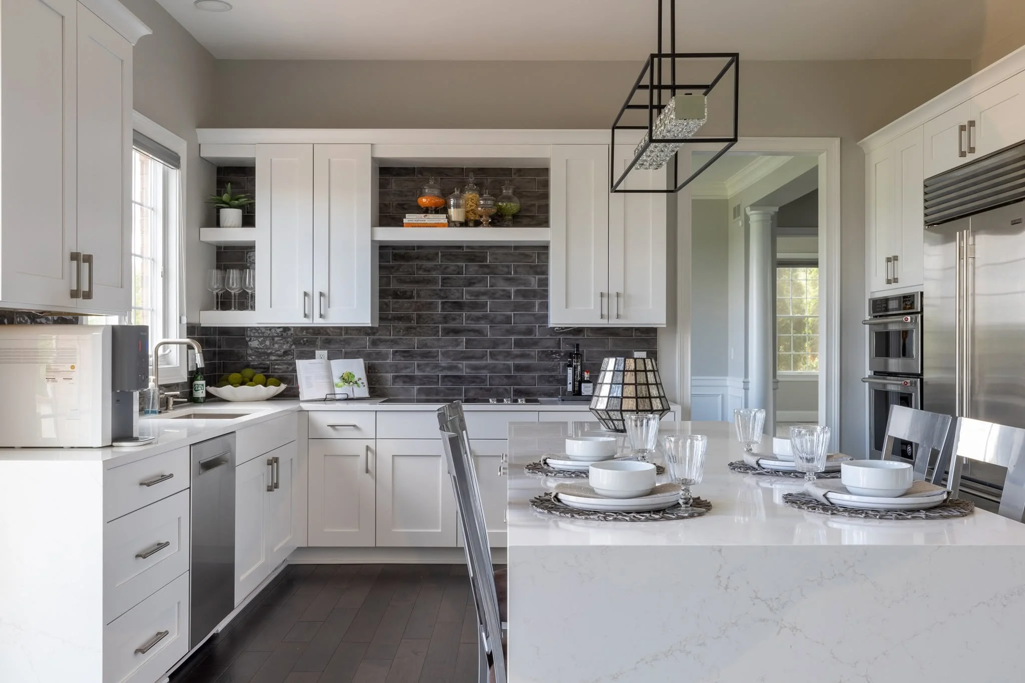 Modern kitchen with white cabinets, dark brick backsplash, a kitchen island with place settings, and a dining table with glasses and bowls.