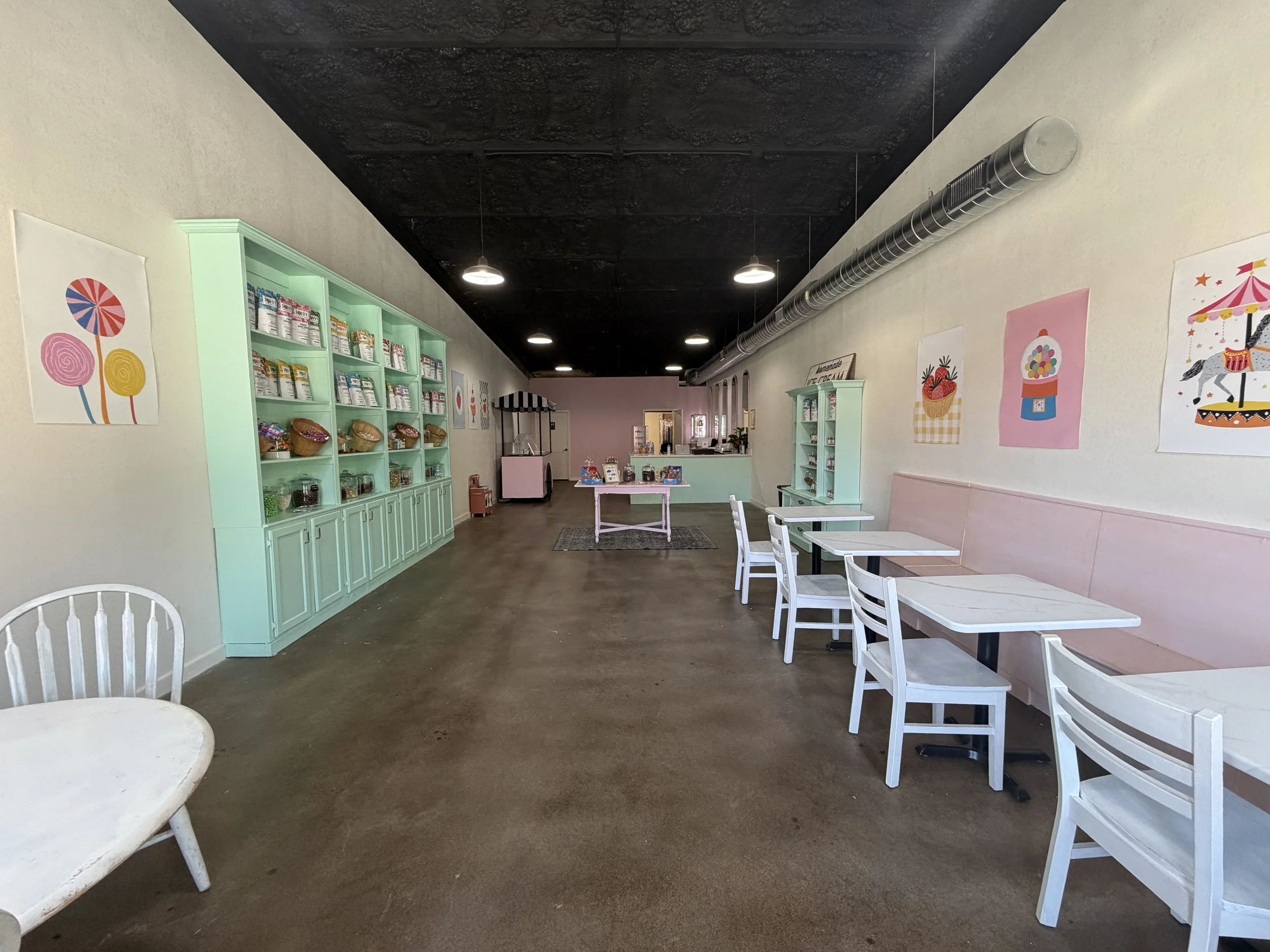 Interior of a pastel-colored ice cream or candy shop with mint green shelves stocked with items, pink walls decorated with colorful art, white tables and chairs, and a black ceiling with exposed ductwork.