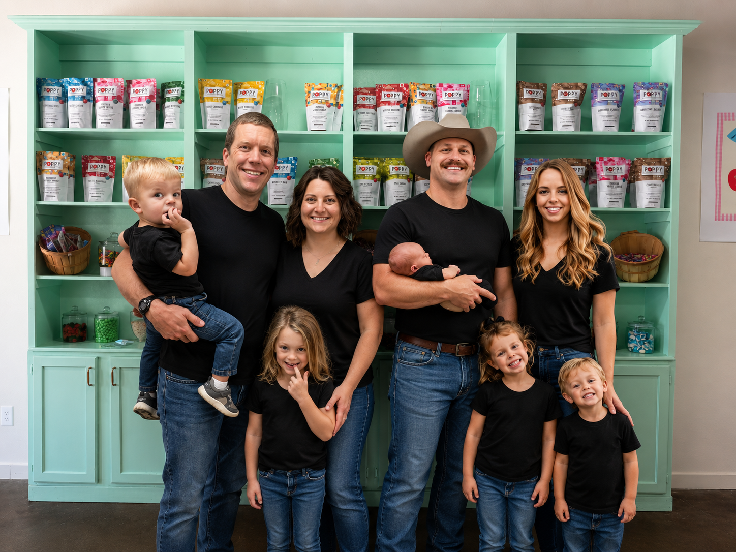 A group of seven people, including four children and three adults, standing in front of a mint green shelf with snack bags. They are smiling and wearing black shirts and jeans. The adults are holding two babies, one on each side. The children are standing in front, with one girl smiling and pointing, and three boys smiling.