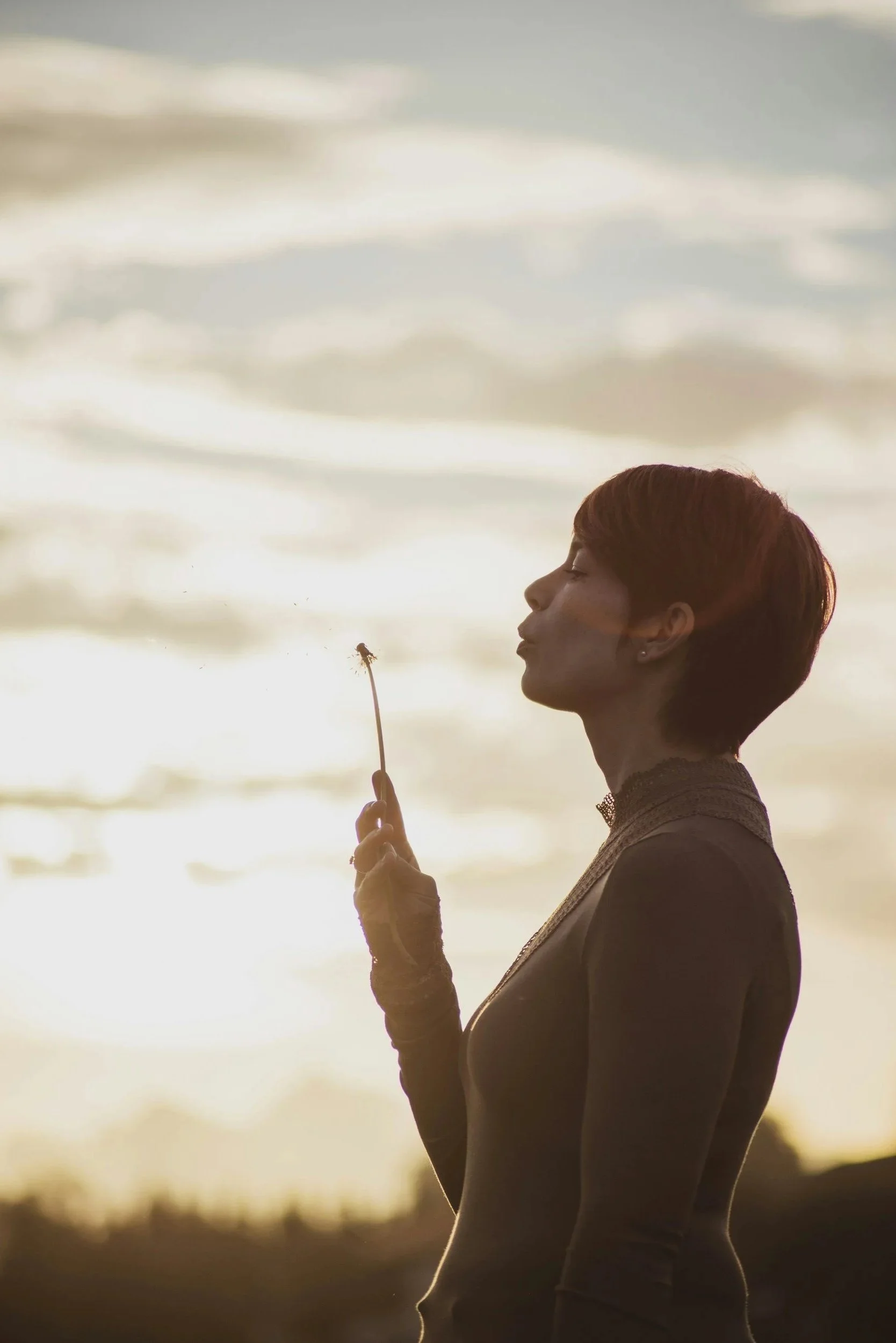 A woman leaving her session at Hopeful Healing Therapy in Kansas City with short hair holding a dandelion in her hand, blowing the seeds into the air, with a cloudy sky in the background during sunset.