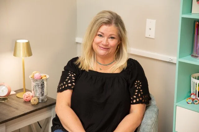 Nikki Hope Fryer, LCSW from Hopeful Healing Therapy in Kansas City with blonde hair smiling, sitting in a room with a gray wall, a gold lamp, and a table with decorative balls and a jar of yarn.