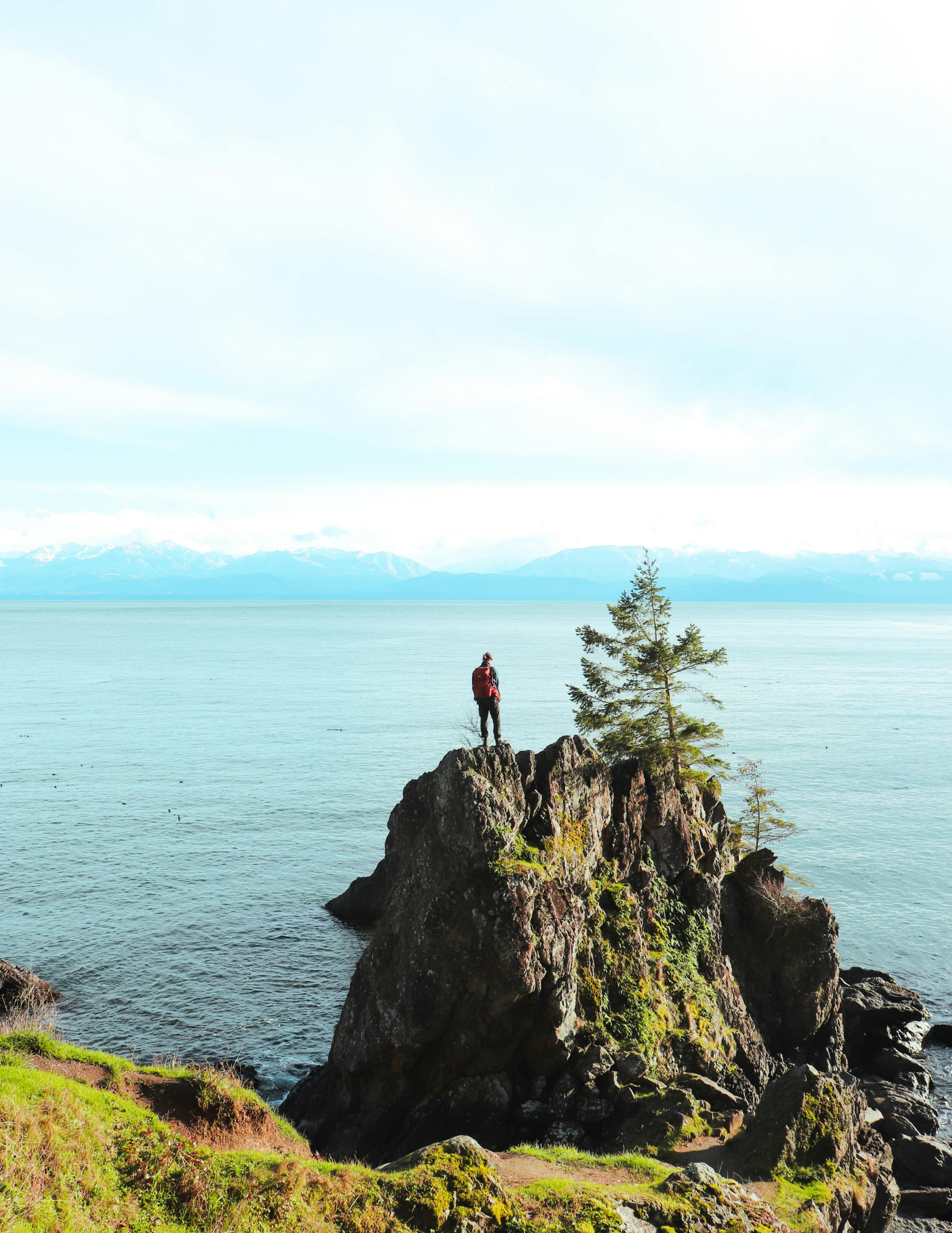 A person with a red backpack standing on rocks on a island overlooking a large body of water with mountains in the distance and a partly cloudy sky.