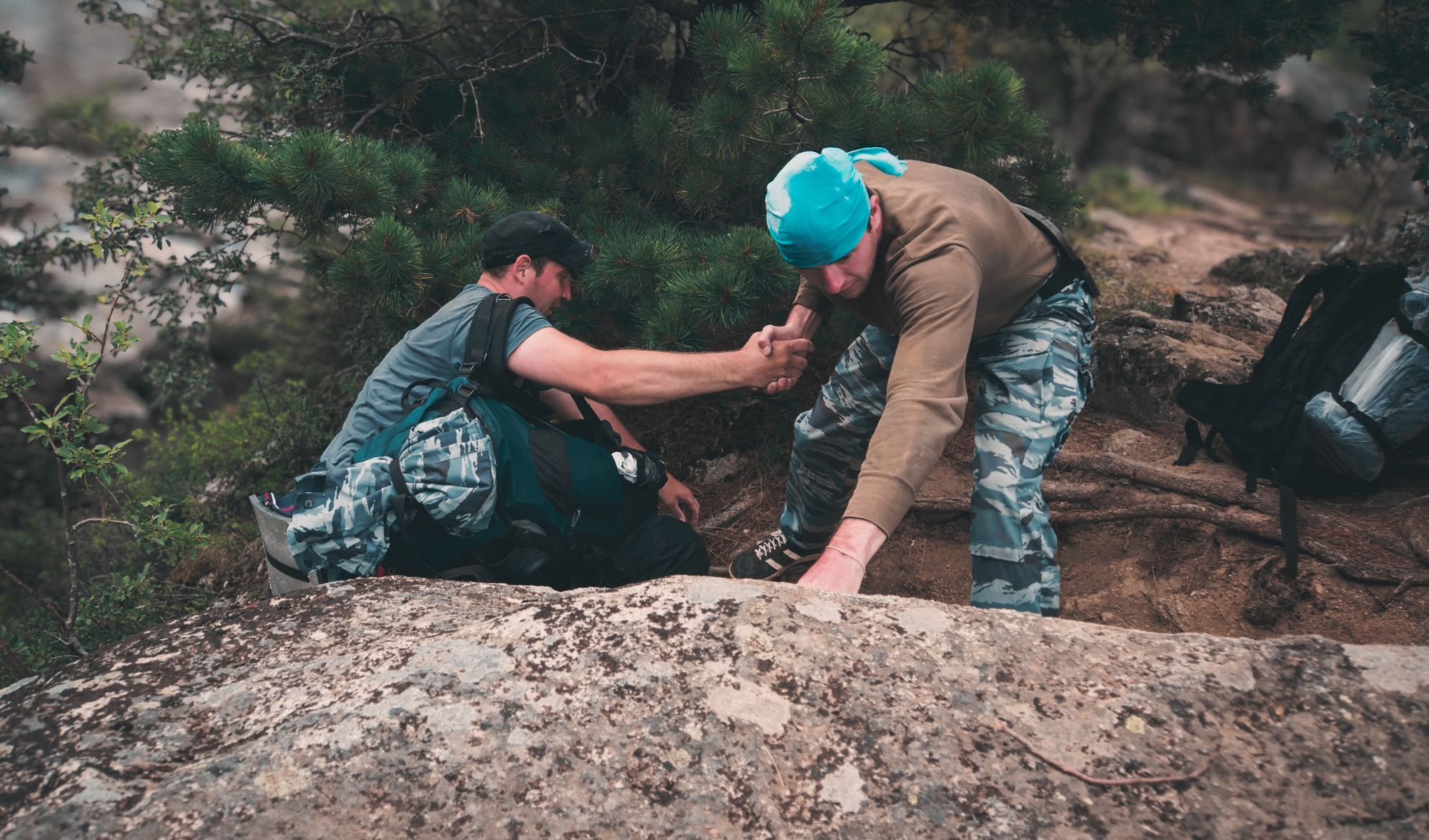 Two men on a mountain trail, one helping the other, both dressed in outdoor gear, with backpacks, near a pine tree and rocky terrain.