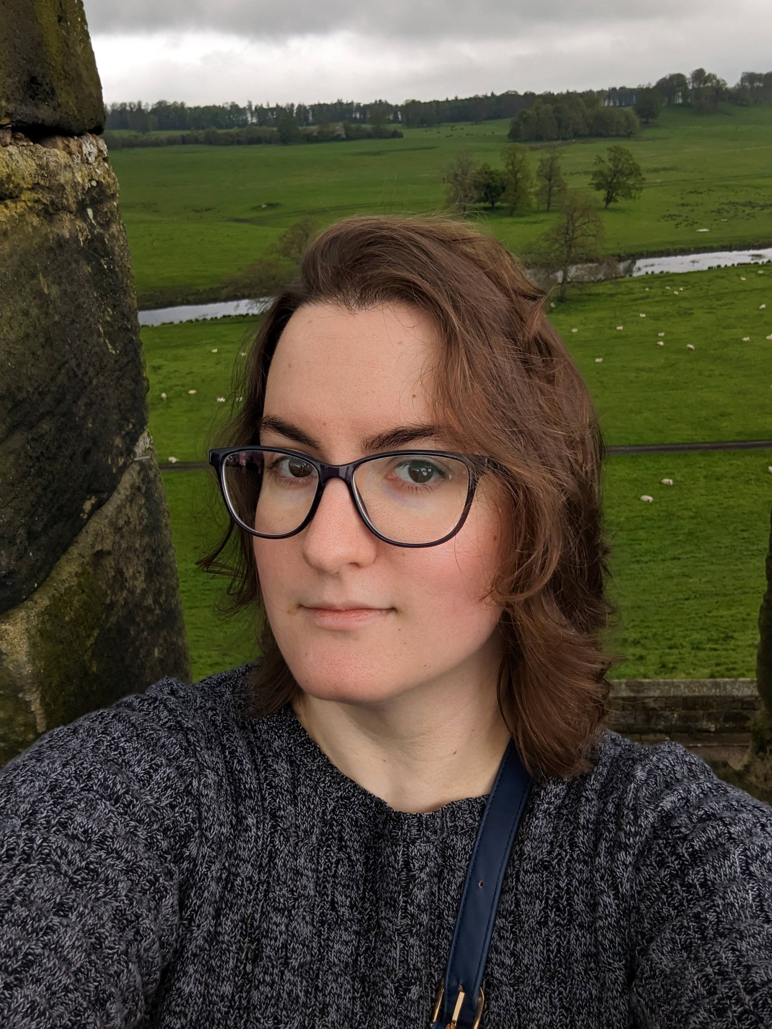 A young woman with brown hair, glasses, and a gray sweater, standing outdoors near a stone structure, with green hills, trees, a river, and sheep in the background under a cloudy sky.